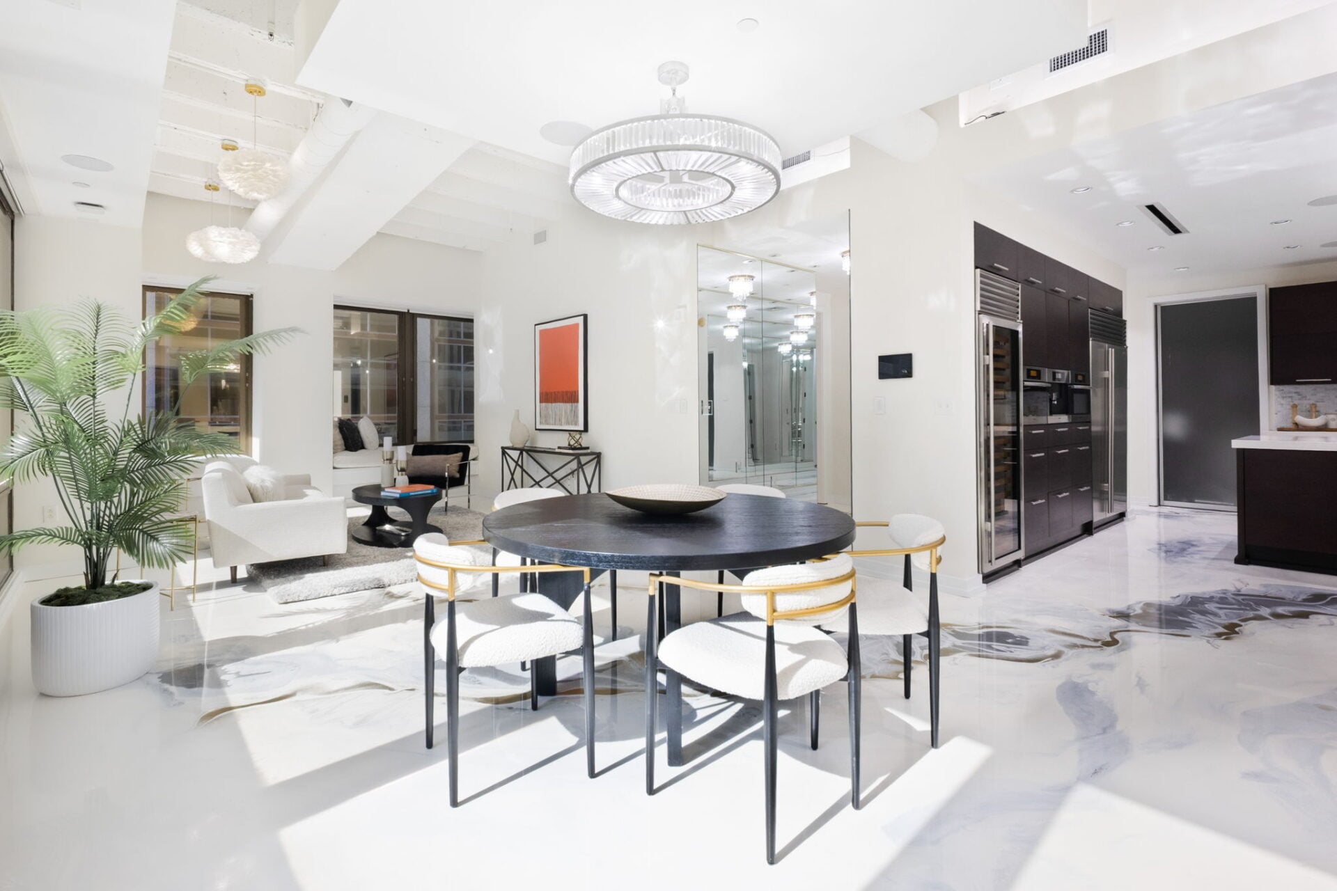 A round dark table is centered beneath a large circular chandelier, with white upholstered chairs arranged around it.