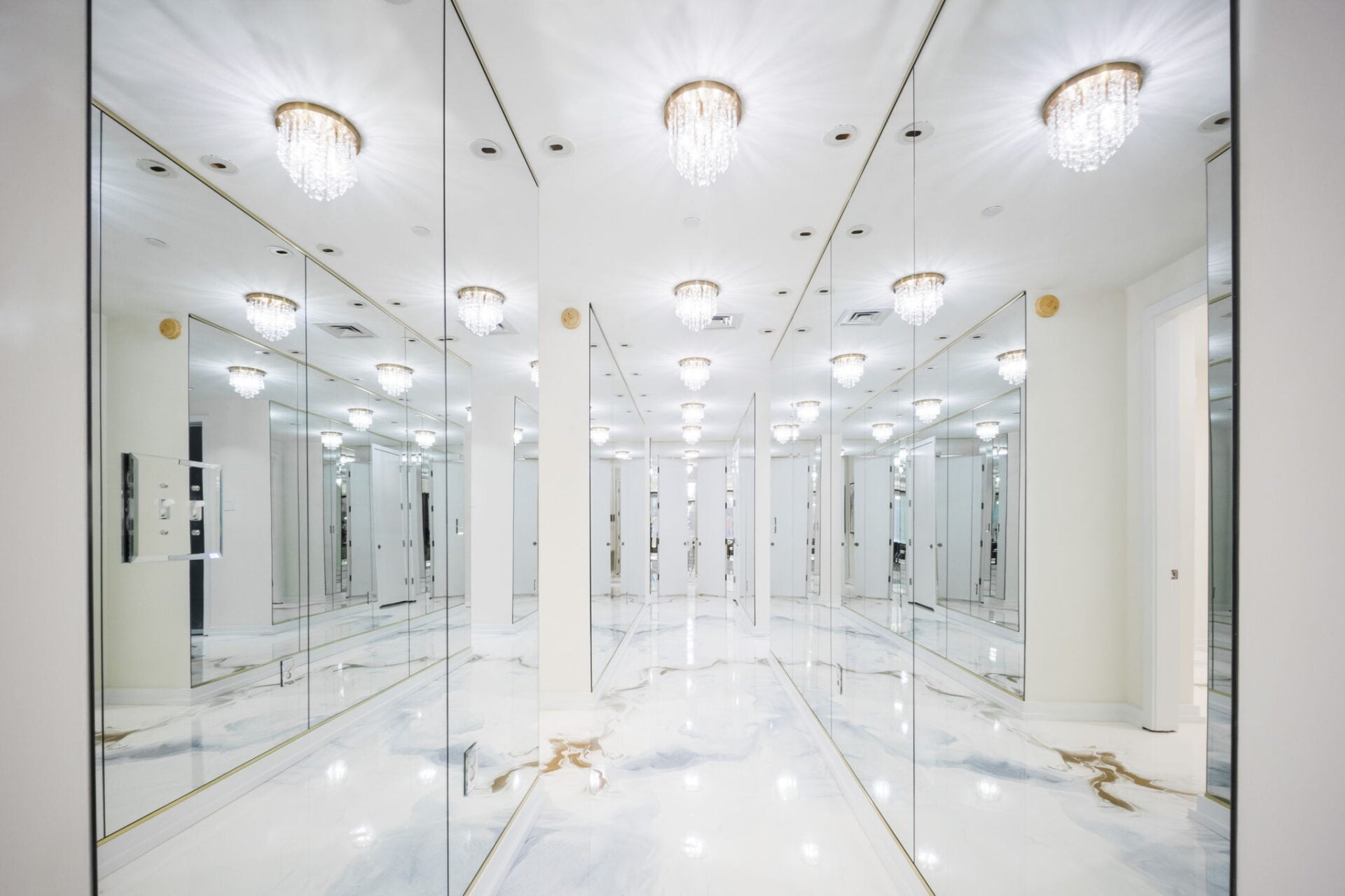 A long mirrored hallway with full-height mirrors lining both walls, reflecting the marble flooring and a series of crystal lights overhead.