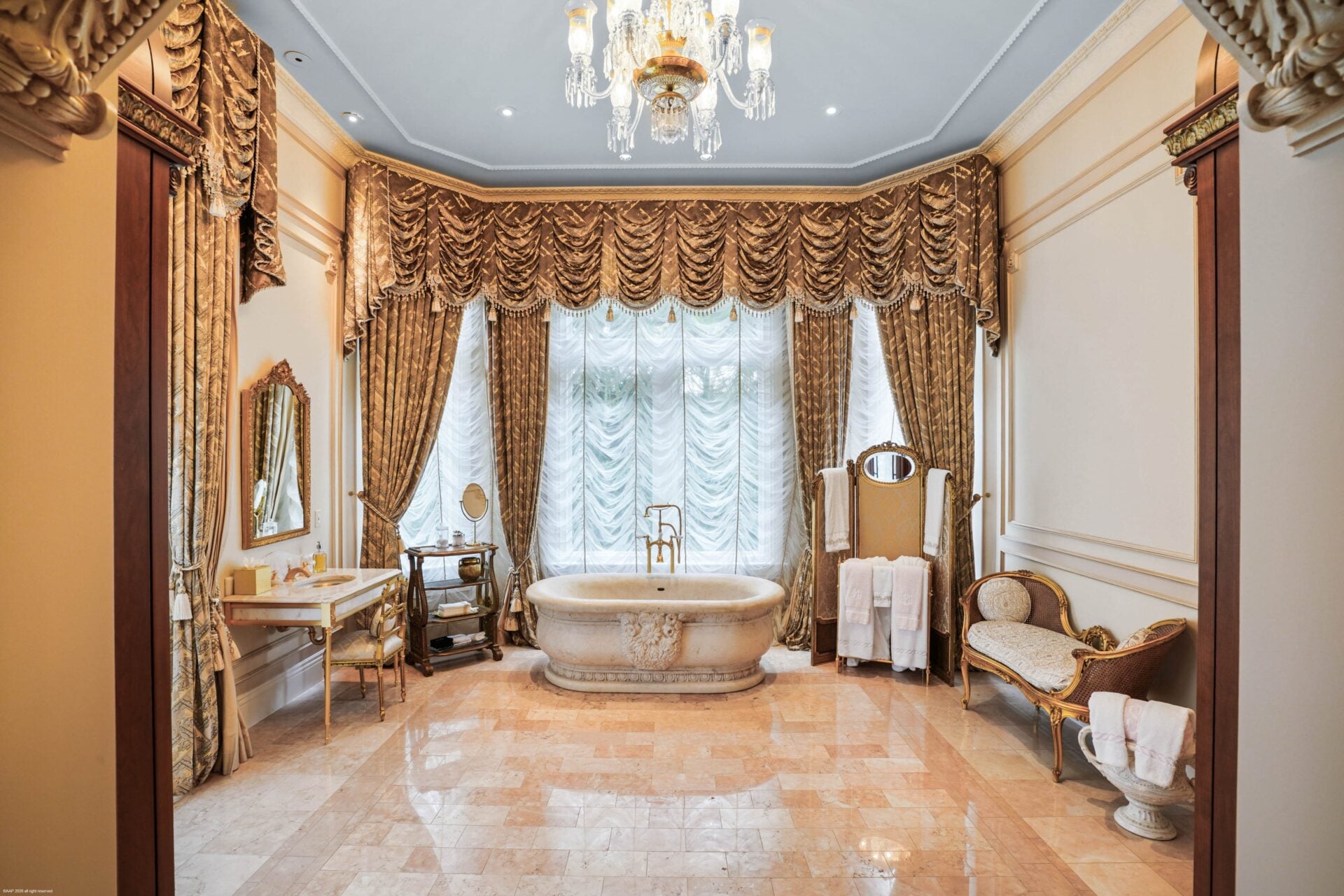 Bathroom with pale stone flooring, a freestanding soaking tub in front of a wide window, and heavy drapery framing the space.