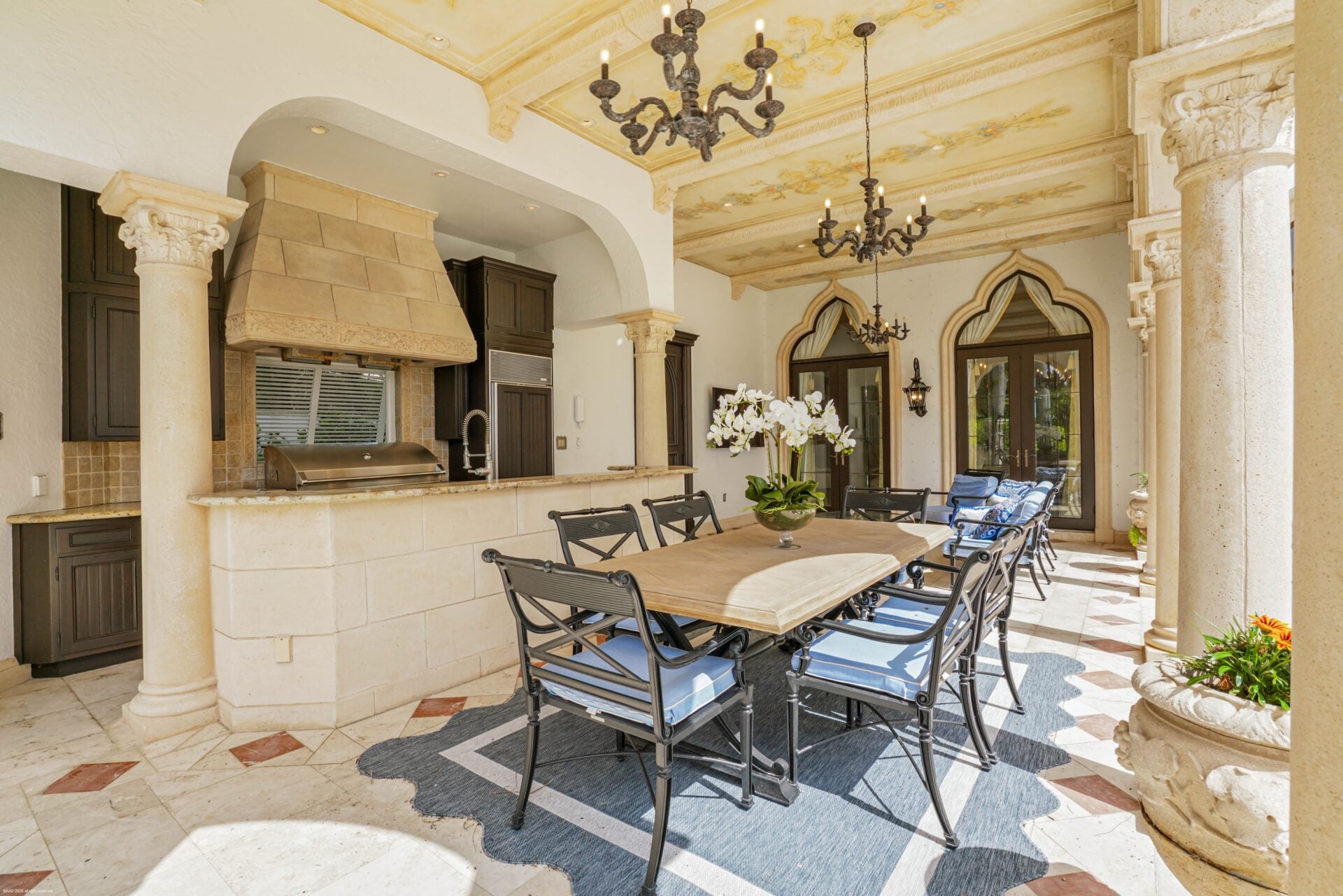 The opposite side has an outdoor kitchen with a built-in grill, sink, lots of dark cabinetry, and a long dining table set beneath the covered roof.