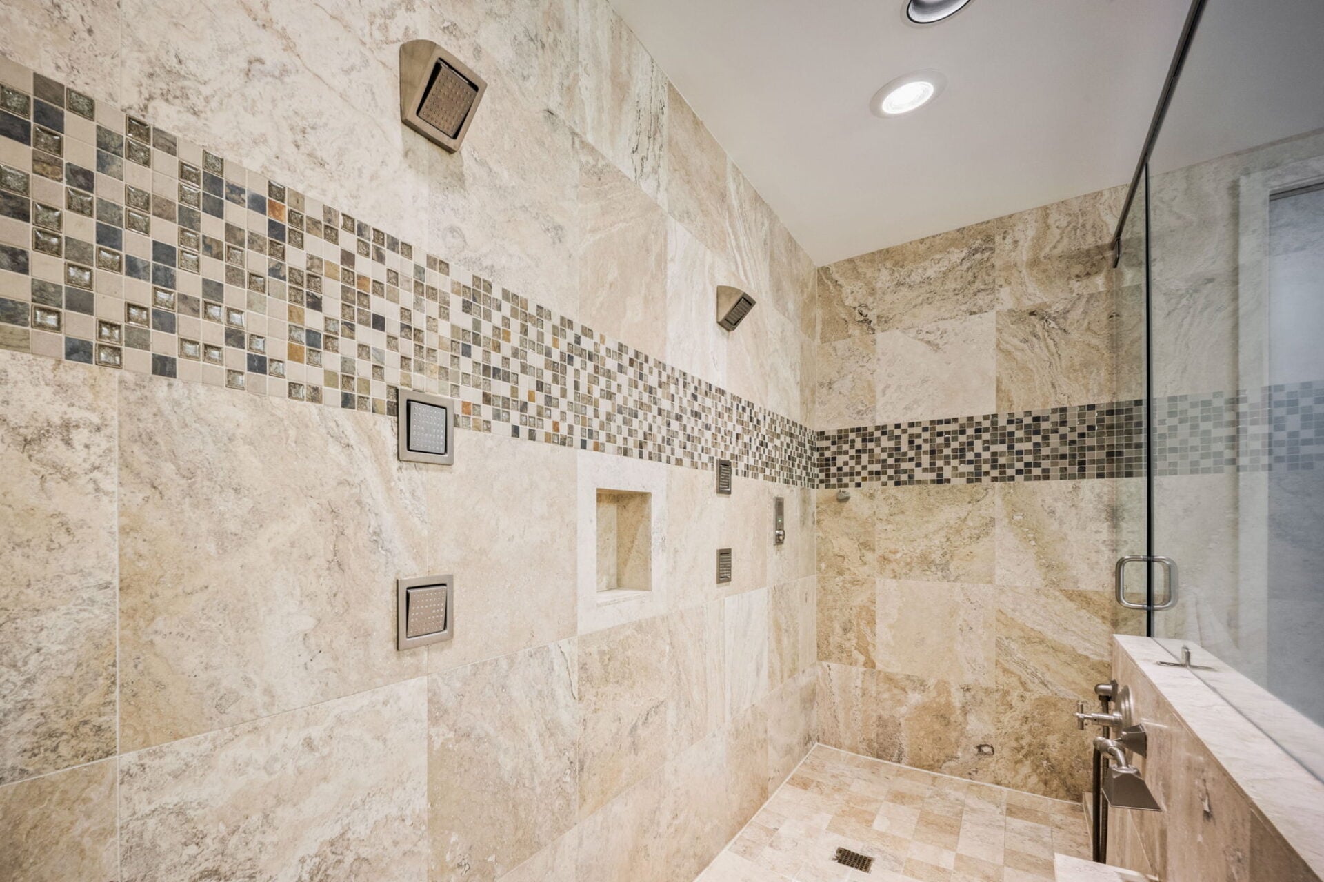 Inside the two-person shower, full-height stone tile is broken by a horizontal mosaic band, with two sets of wall-mounted jets and overhead fixtures spread out along the back wall.