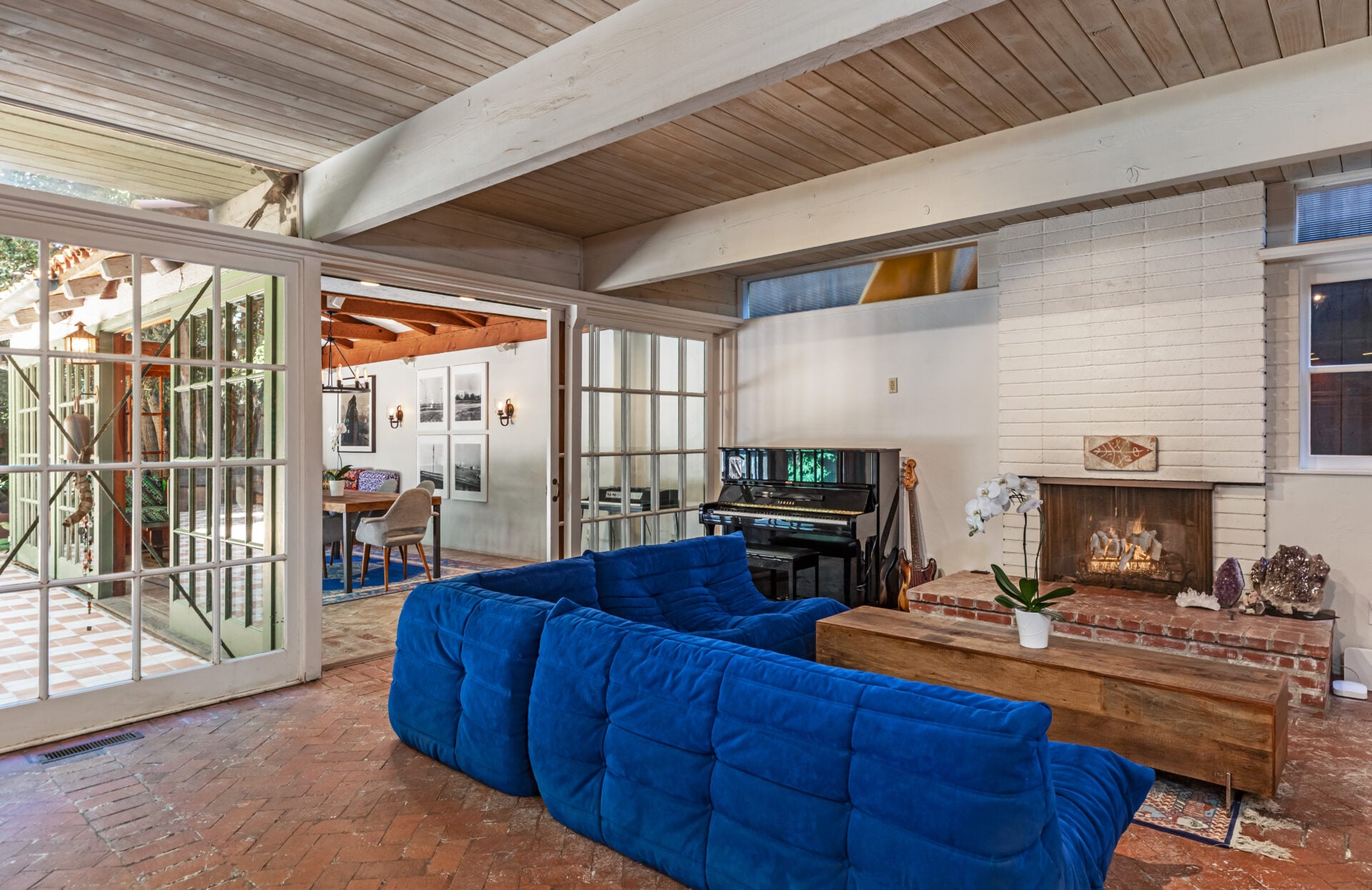 The living area gathers beneath the exposed ceiling beams around a brick fireplace, with a deep blue couch positioned directly in front of it. 