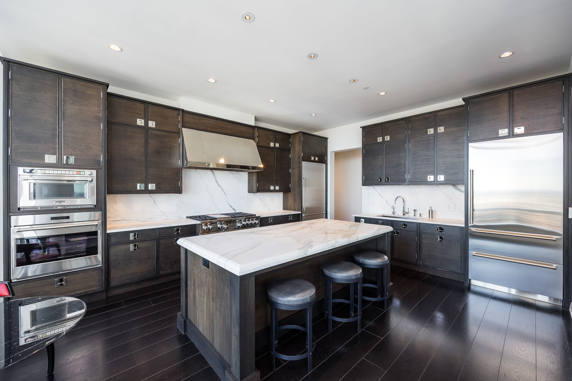 the kitchen is wrapped in custom dark wood cabinetry lining the walls around the stainless-steel appliances and a large stone-topped island at the center.
