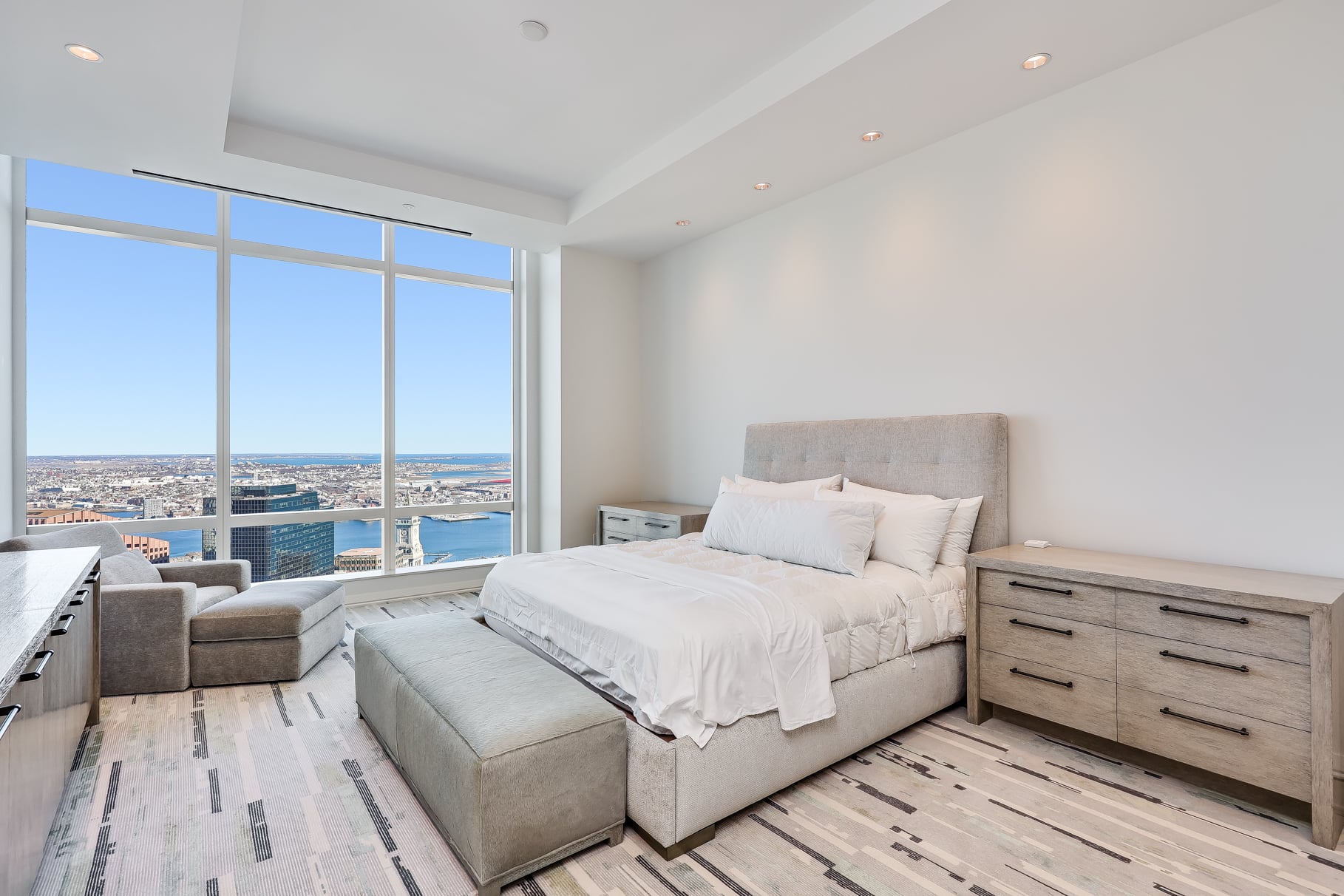 A lounge chair and ottoman sit near the glass, while dressers sit on either side of the bed.