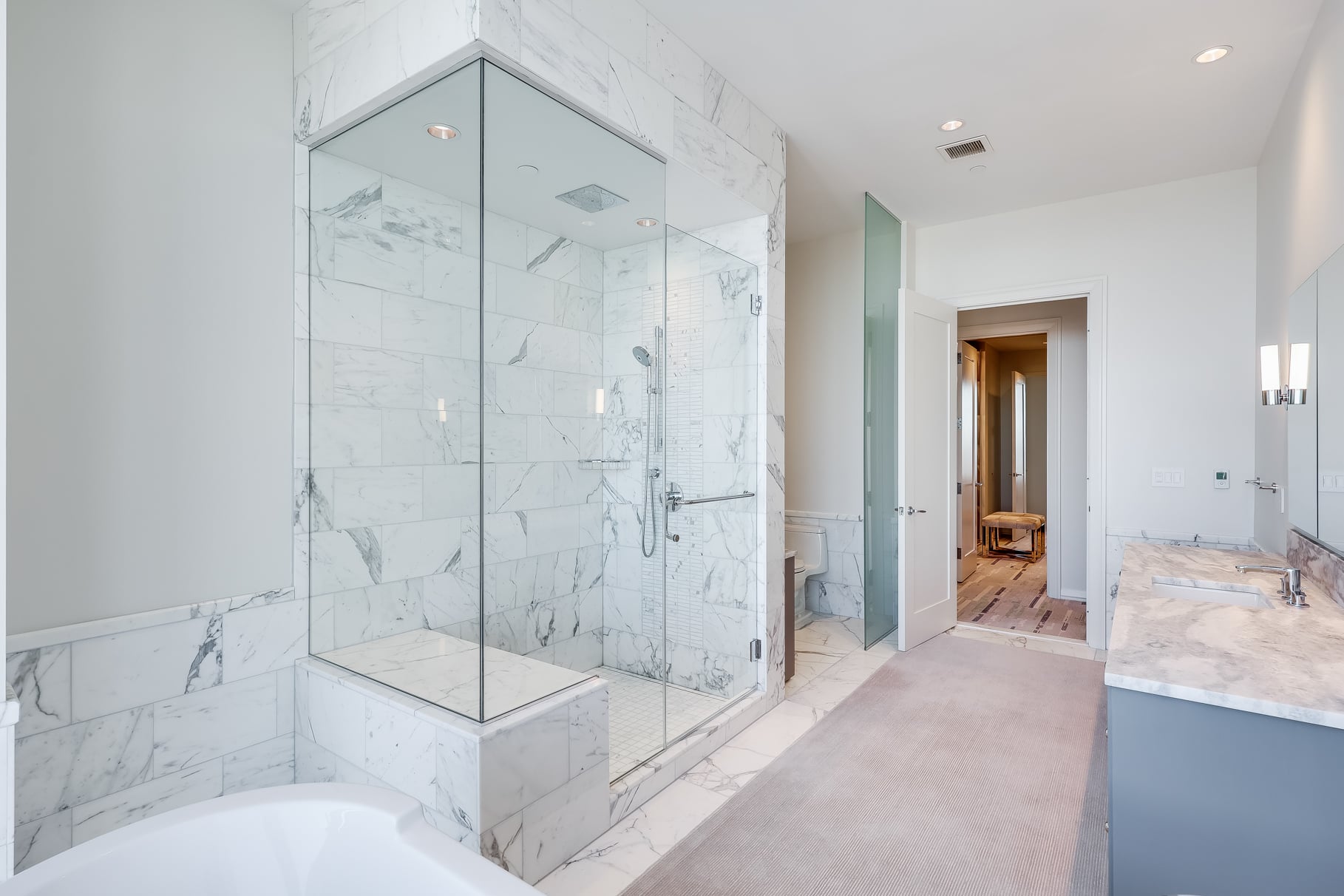 Across from both the vanity and tub, a large glass-enclosed shower is wrapped in matching marble and includes a built-in bench.