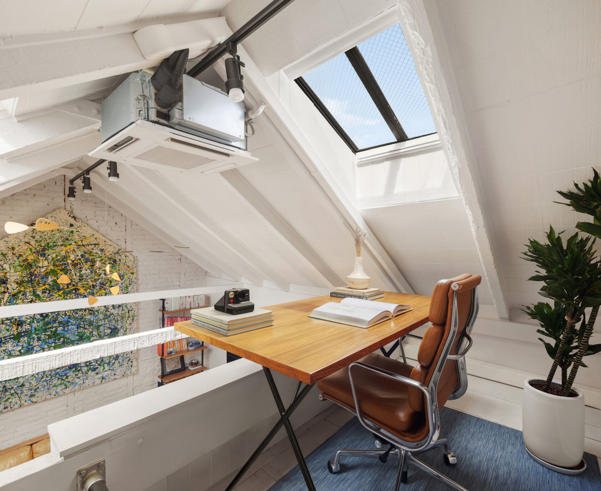 An open loft overlooks the main living area from beneath another skylight, with the desk placed along the railing so the workspace looks directly down into the room below.