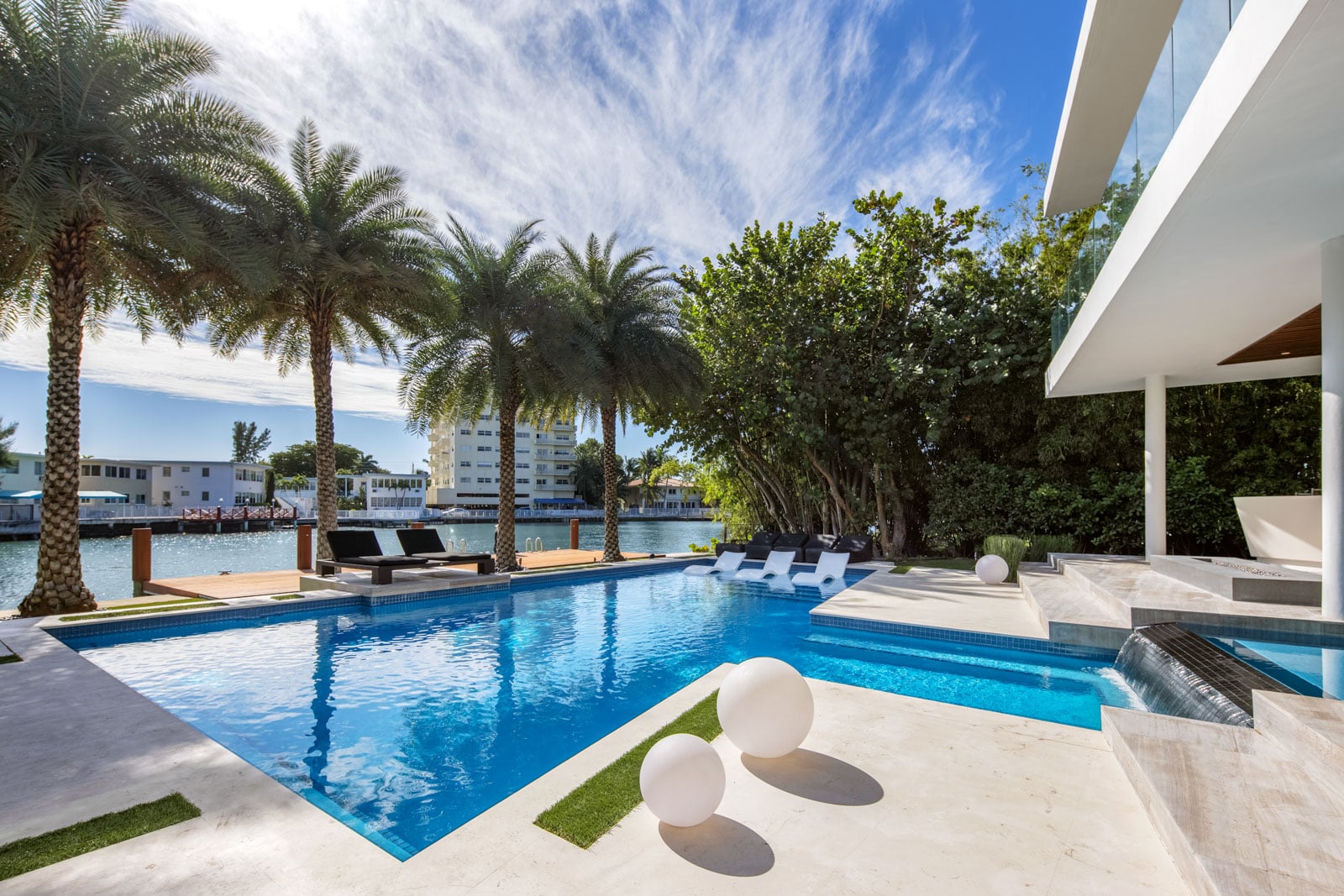 The pool extends toward the property’s roughly 90 feet of waterfront, framed by palm trees, in-water loungers, and additional seating along the edge.