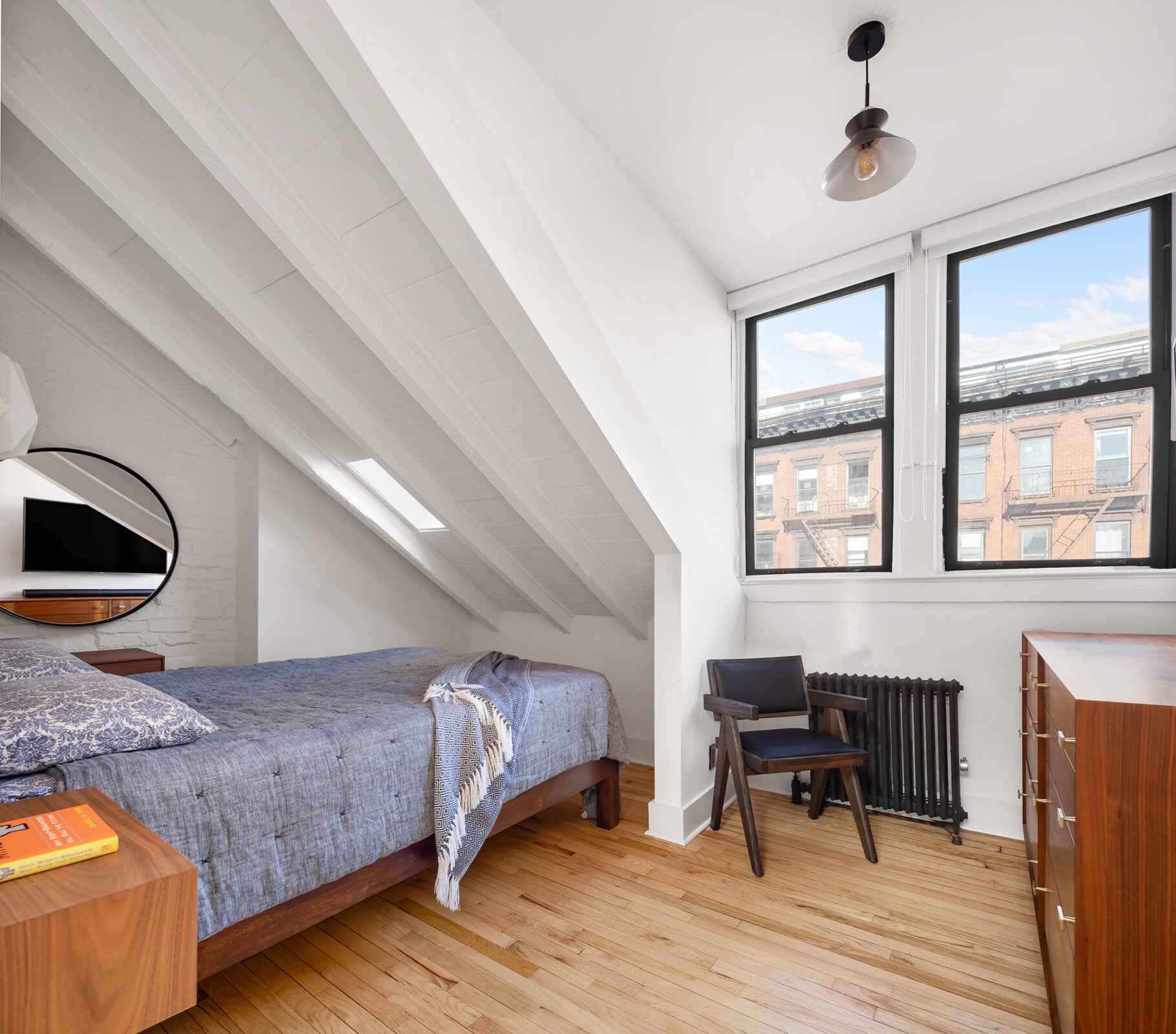 A narrow skylight cuts into the angled ceiling above the bed, while two larger windows at the opposite end look out over the surrounding streets and nearby treetops.