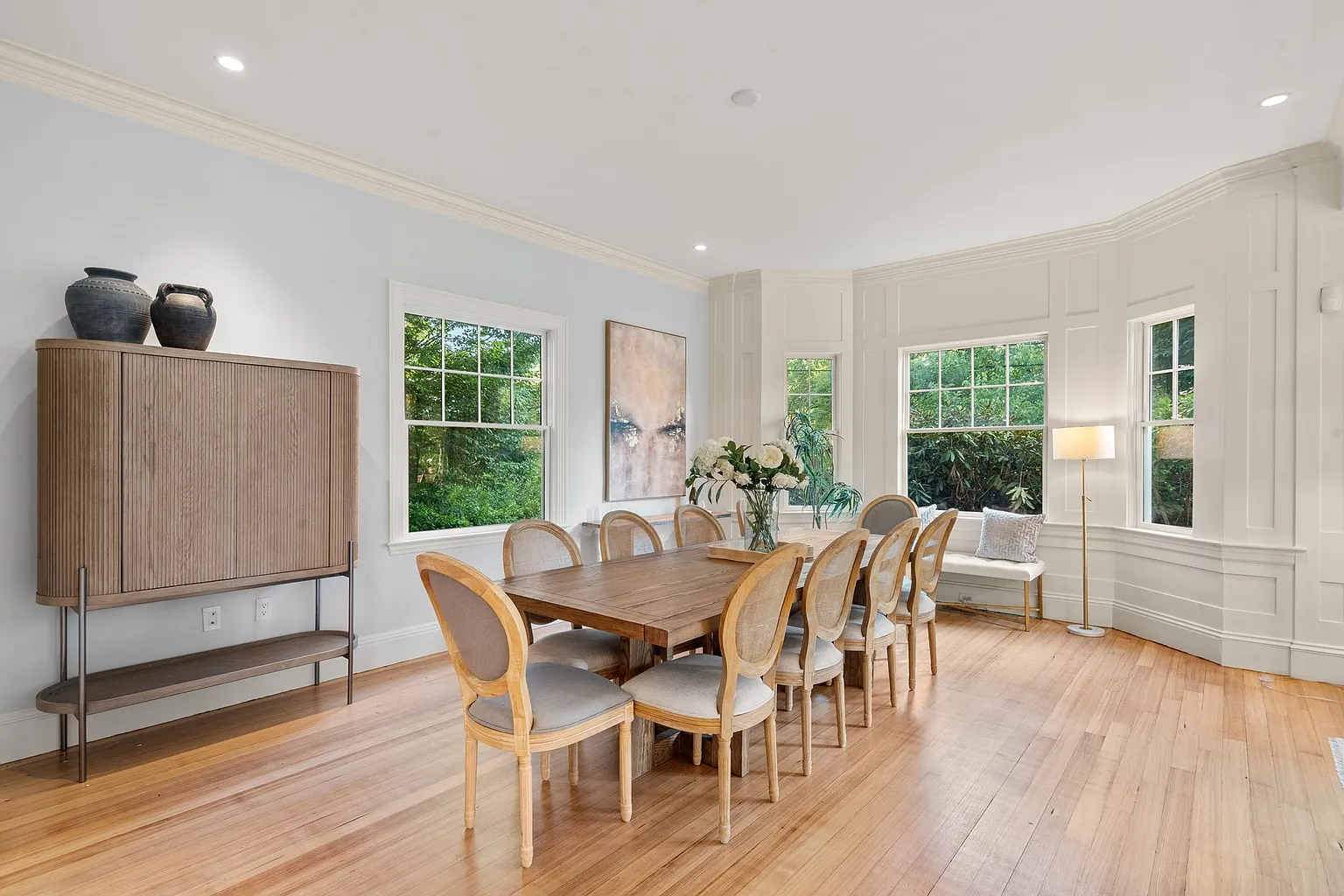 The dining area is palced at the back of the room with a long table centered between multiple windows and paneled walls.