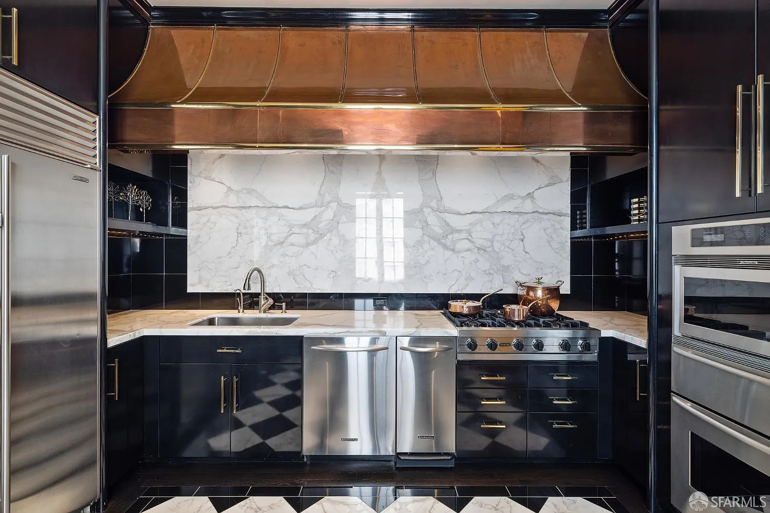 An oversized copper hood stretches nearly the width of the cooking wall, creating a warm metallic contrast against the black cabinetry and pale stone backsplash.