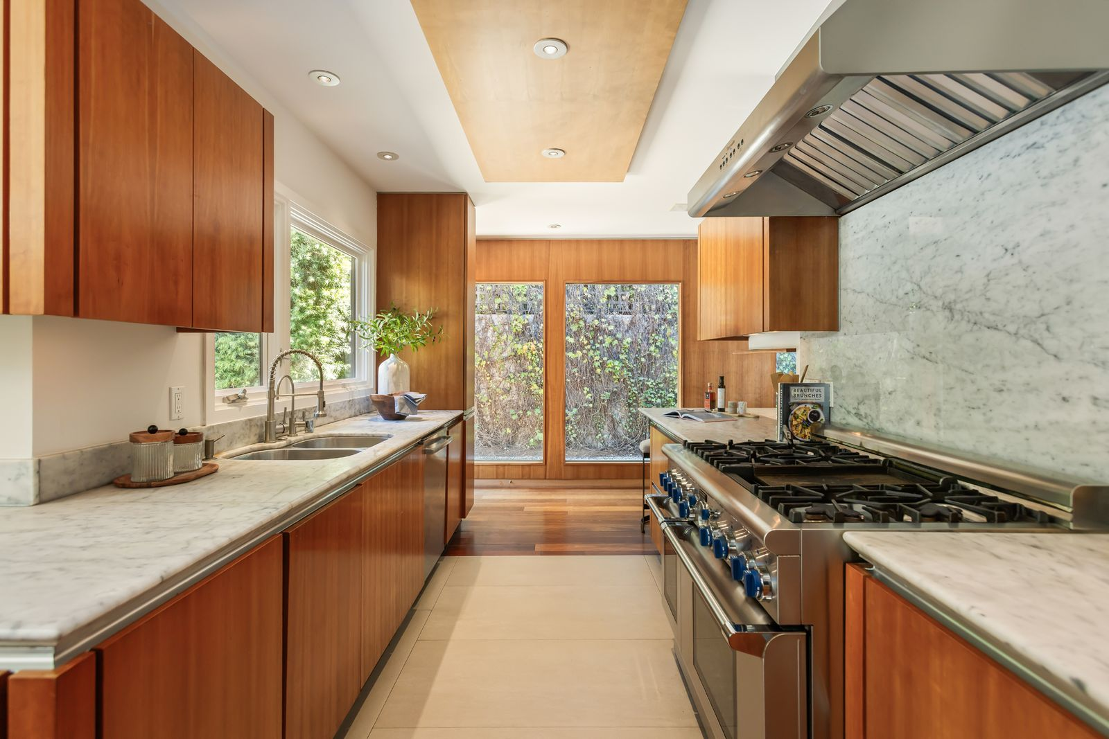 A professional-grade range sits beneath an oversized stainless hood on one side, while the sink and additional prep space run beneath a large window across from it.