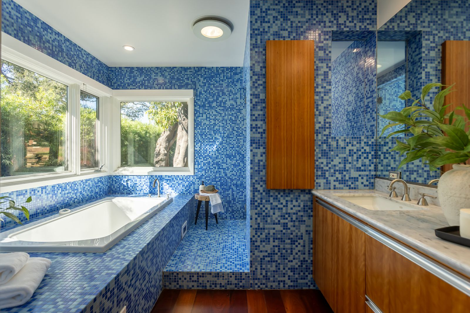 The soaking tub is built into a tiled platform beneath a pair of corner windows, while the vanity runs along the opposite side with wood cabinetry and a marble counter.