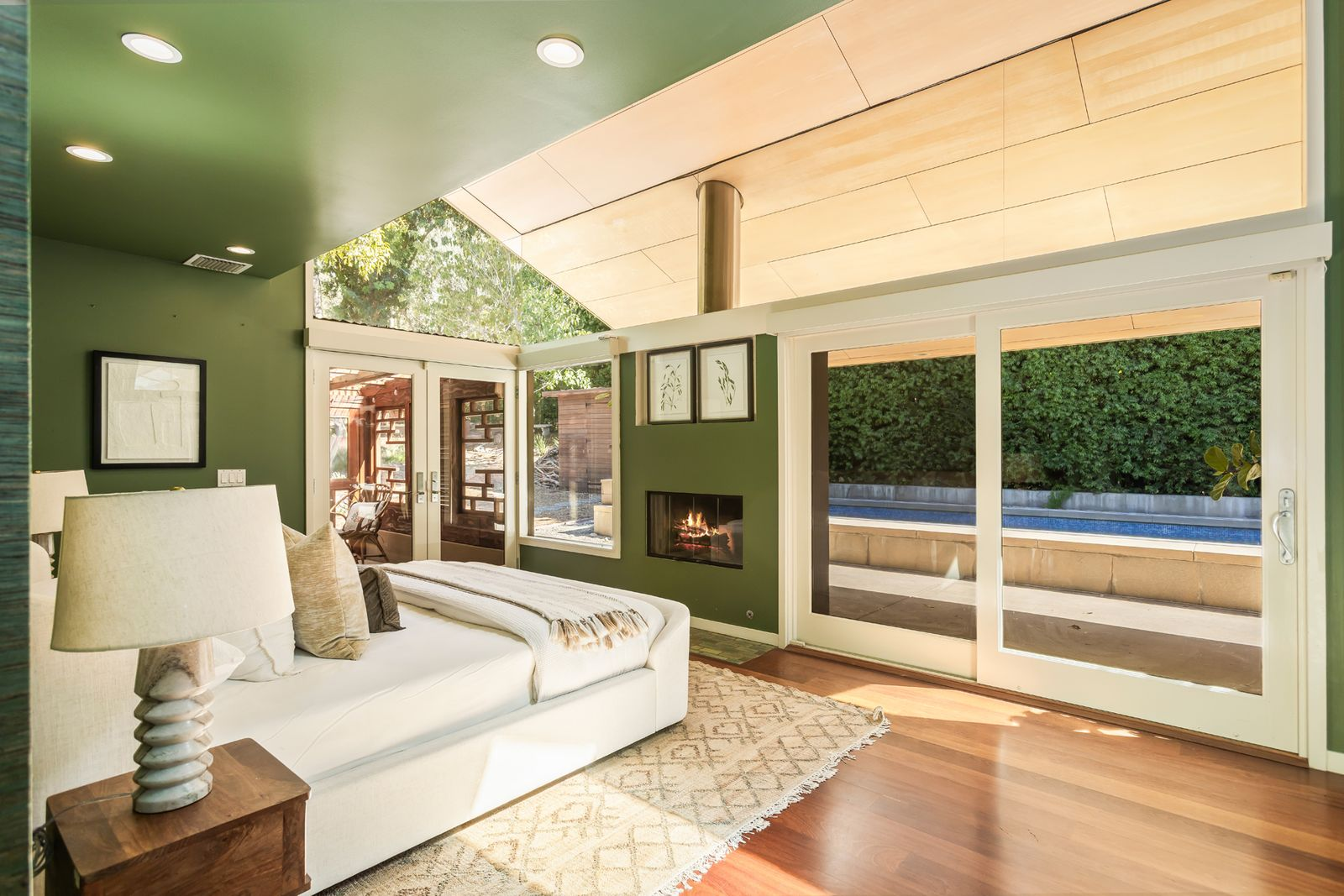 The primary suite opens beneath a vaulted ceiling, with the bed placed across from a fireplace framed by a large glass window on one side and sliding doors opening to the pool on the other.