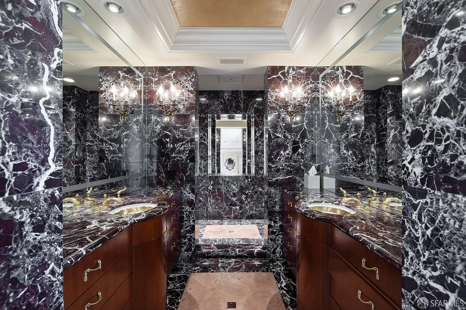 Two separate vanities run along opposite sides of the room before the shower, each with veined dark marble countertops, wood cabinetry, and brass fixtures. 