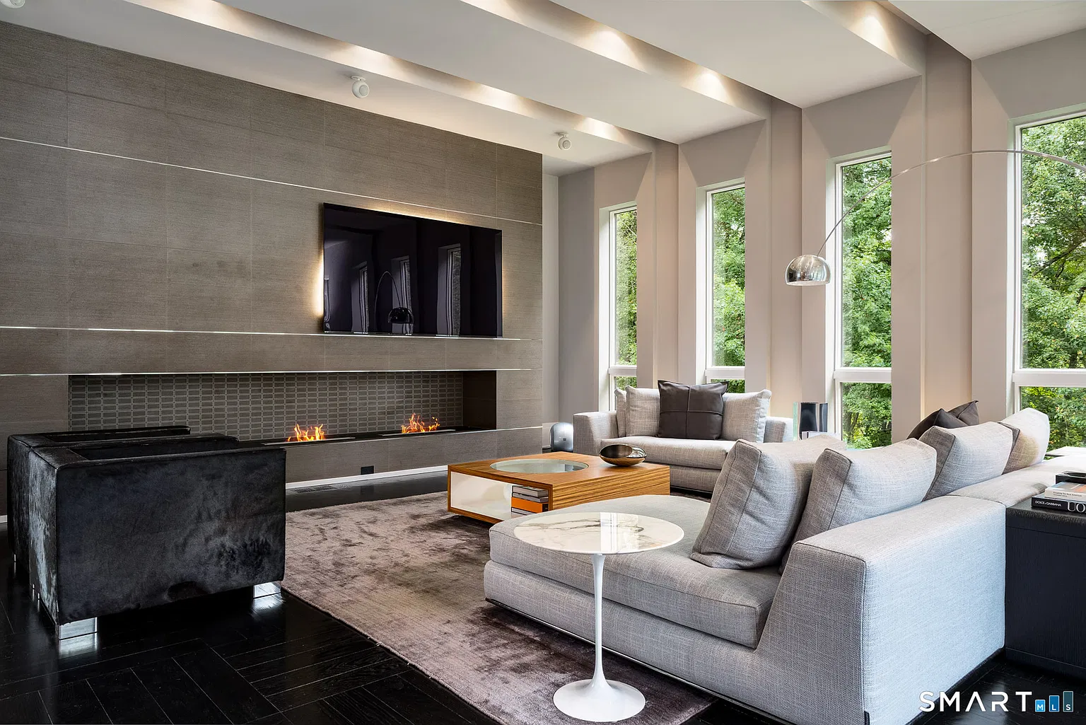 Along one side, the seating arrangement faces a long fireplace built into a full-height feature wall with a mounted television above it. 