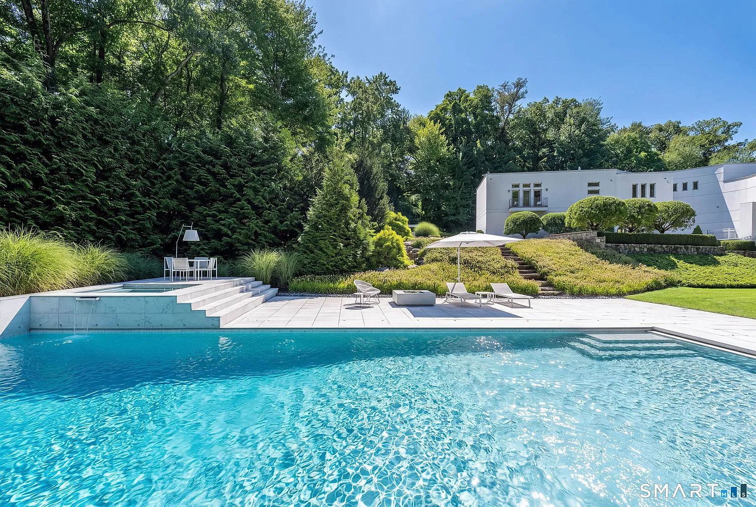 The rectangular pool stretches across the lower grounds, with the raised spa set into one end and separate lounging and dining areas placed along the surrounding deck.