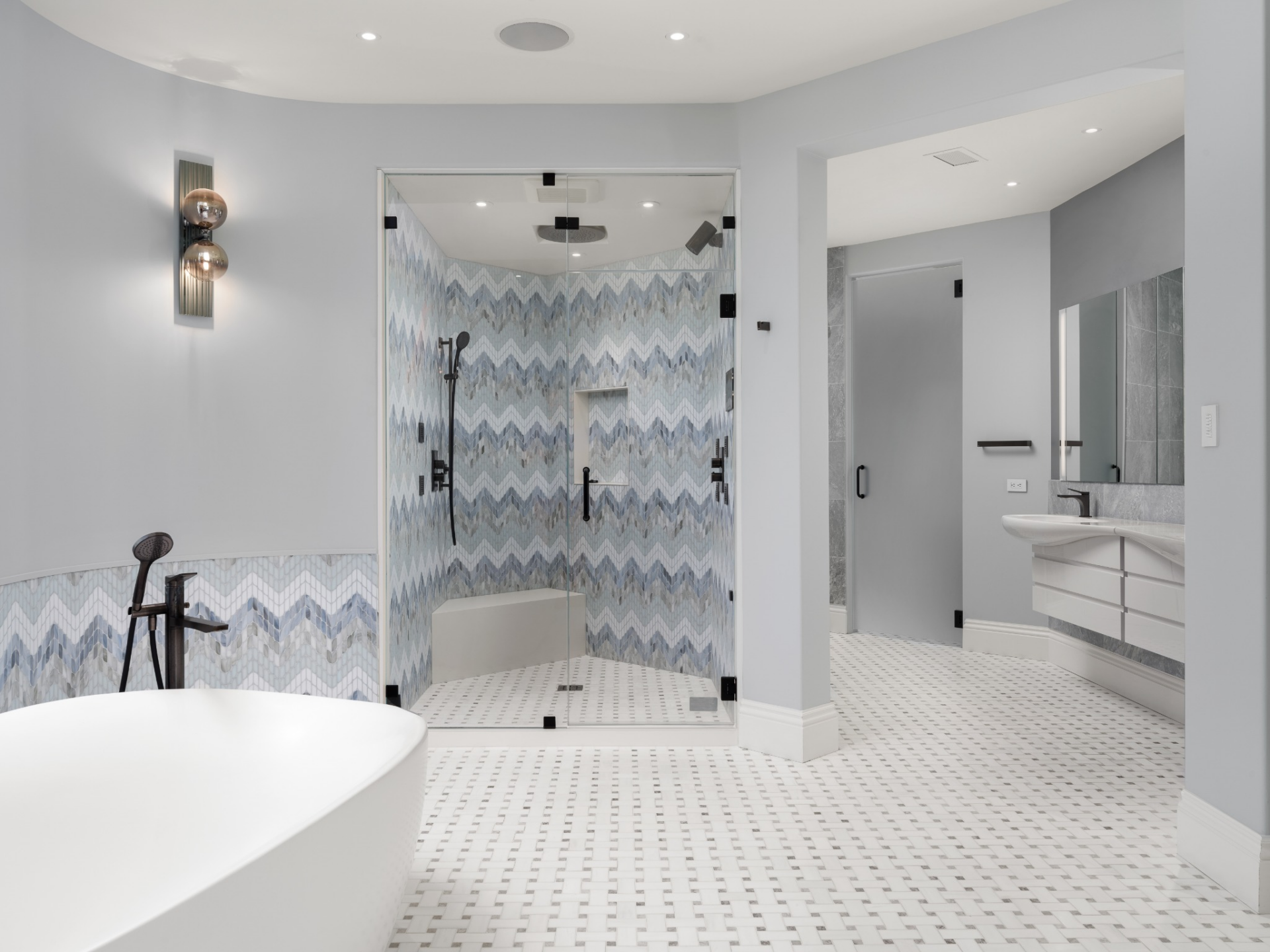 Chevron-patterned tile wrapping the walls of a glass-enclosed shower and moves to the lower portion of the room's wall around a freestanding soaking tub.