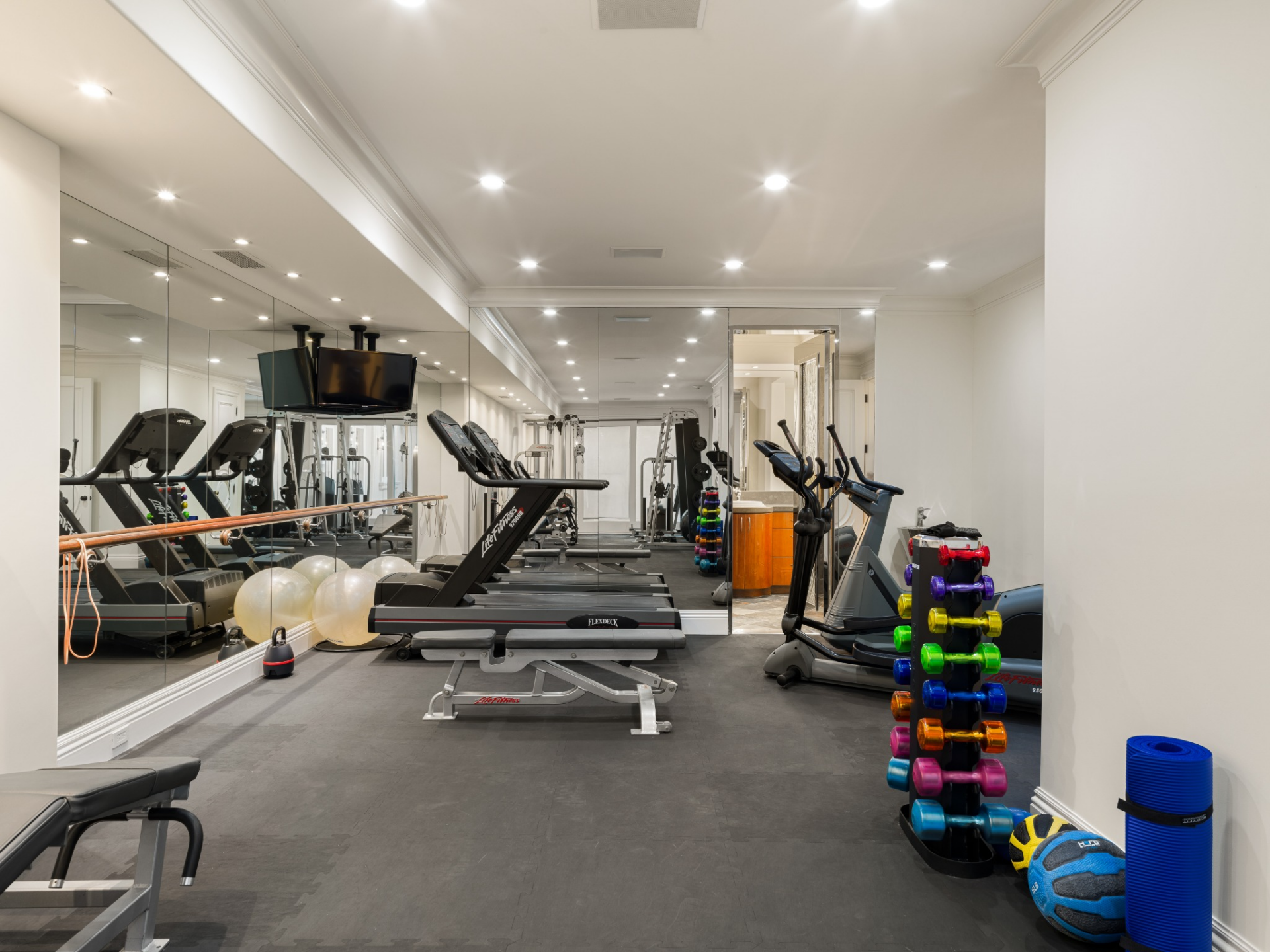The long workout room is lined with mirrored walls and organized around cardio machines, free weights, strength equipment.