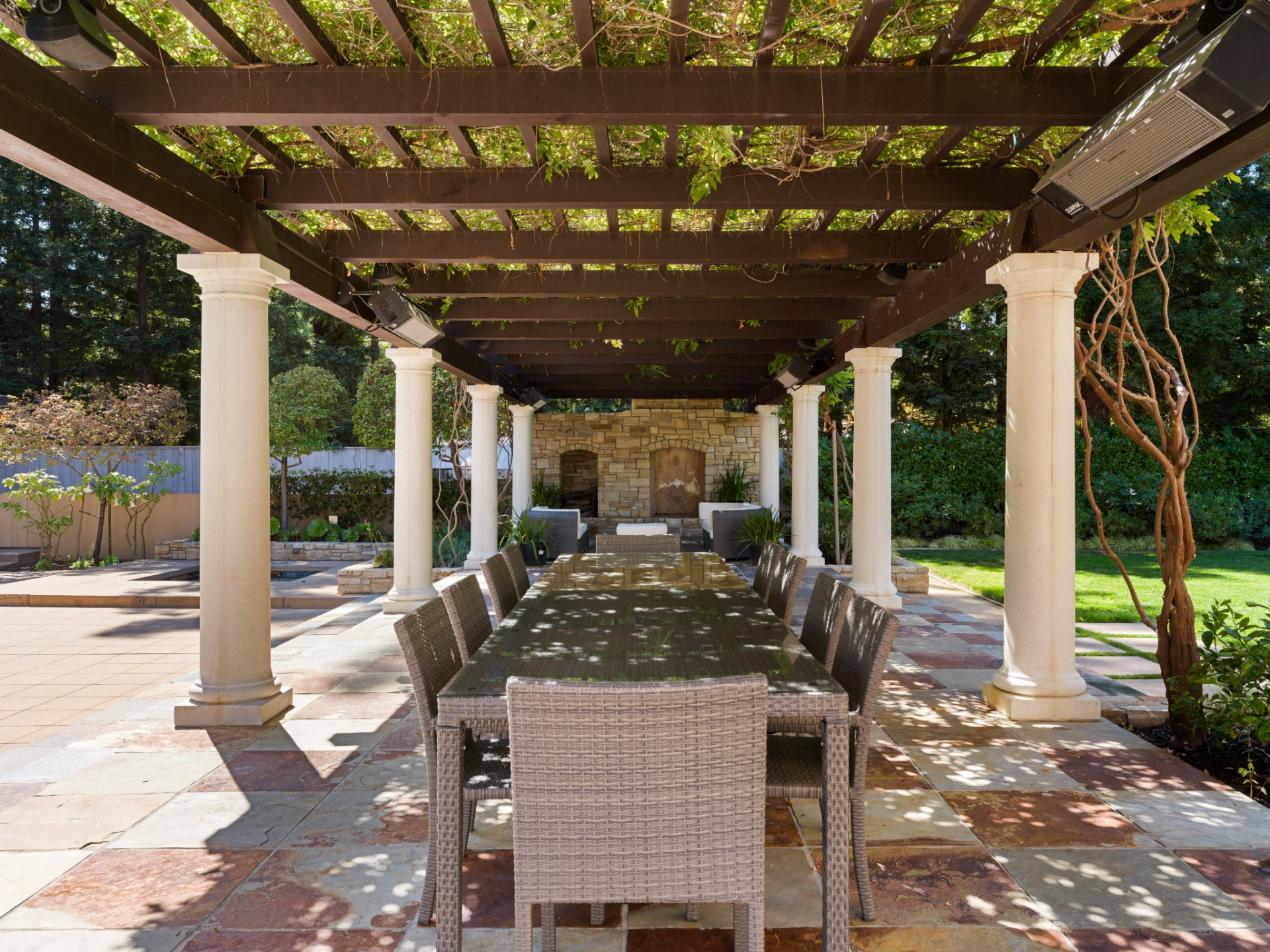 Underneath the vine-covered canopy, one end is set up for outdoor dinners, and the other has a lounge gathered around a stone fireplace.