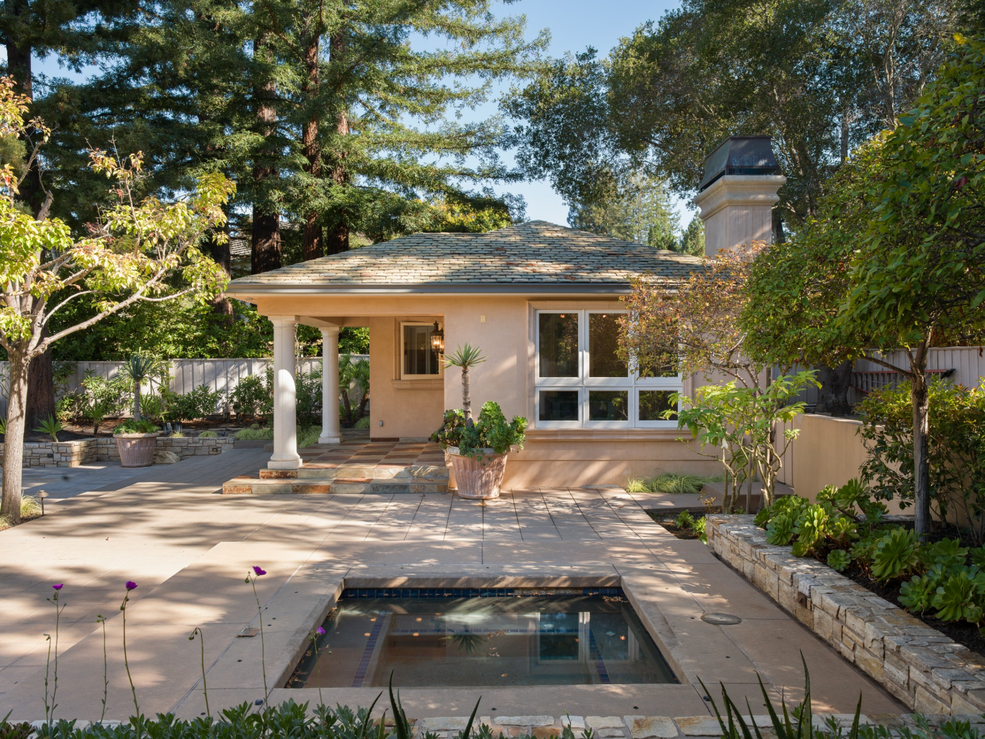 The yard opens to the spa sitting between plant beds and paved terraces with the detached guesthouse across from it.