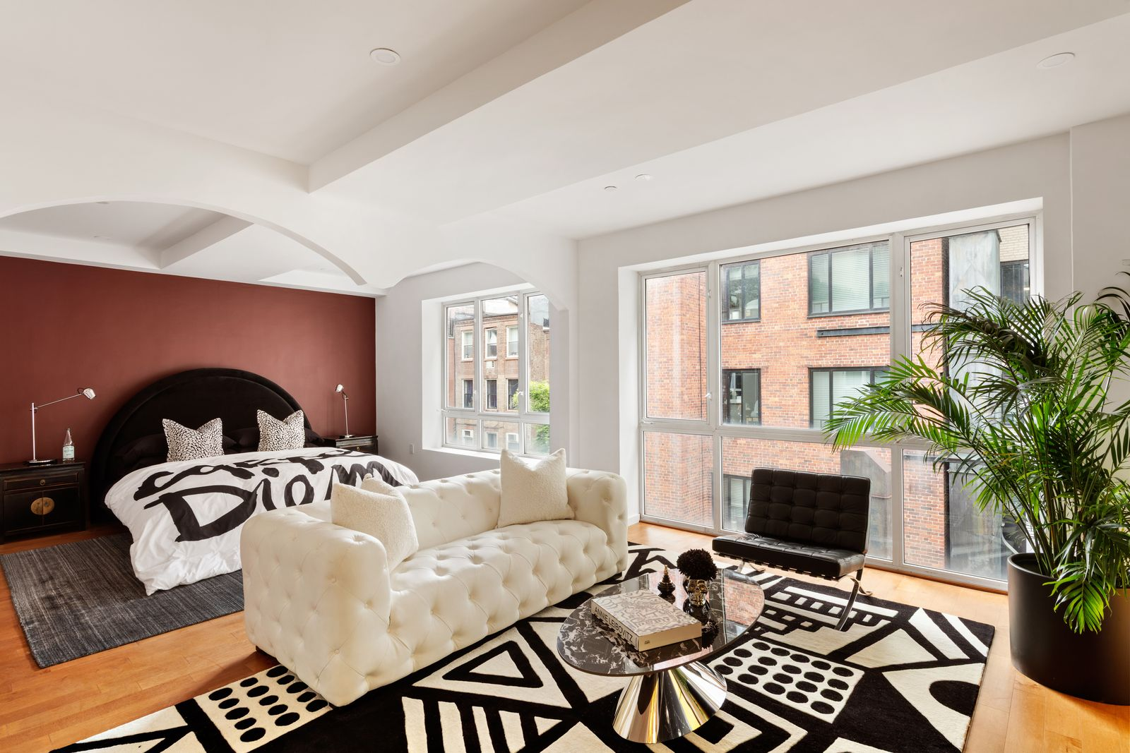 At the back of the room, a dark upholstered bed sits against a deep red accent wall, while a tufted sofa sits in front of it to create a small lounge.
