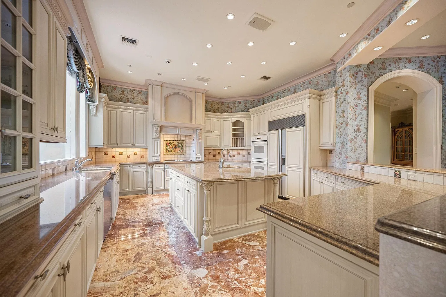 The home’s main kitchen features cream-colored cabinetry lining the flower-patterned walls around a large center island and granite countertops.