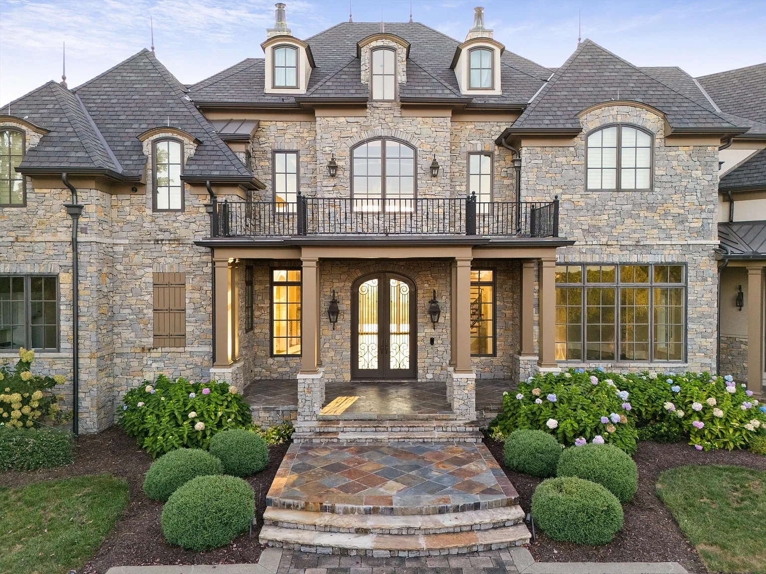 A short set of stone steps rises to the formal entrance, where arched double doors sit beneath a balcony and are framed by matching gas lanterns.
