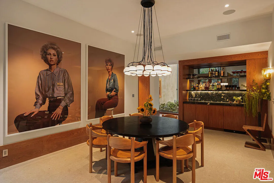 A round table beneath a low-hanging chandelier made up of clustered white glass bulbs, with large-scale artwork filling the wall beside it and a bar at the back.