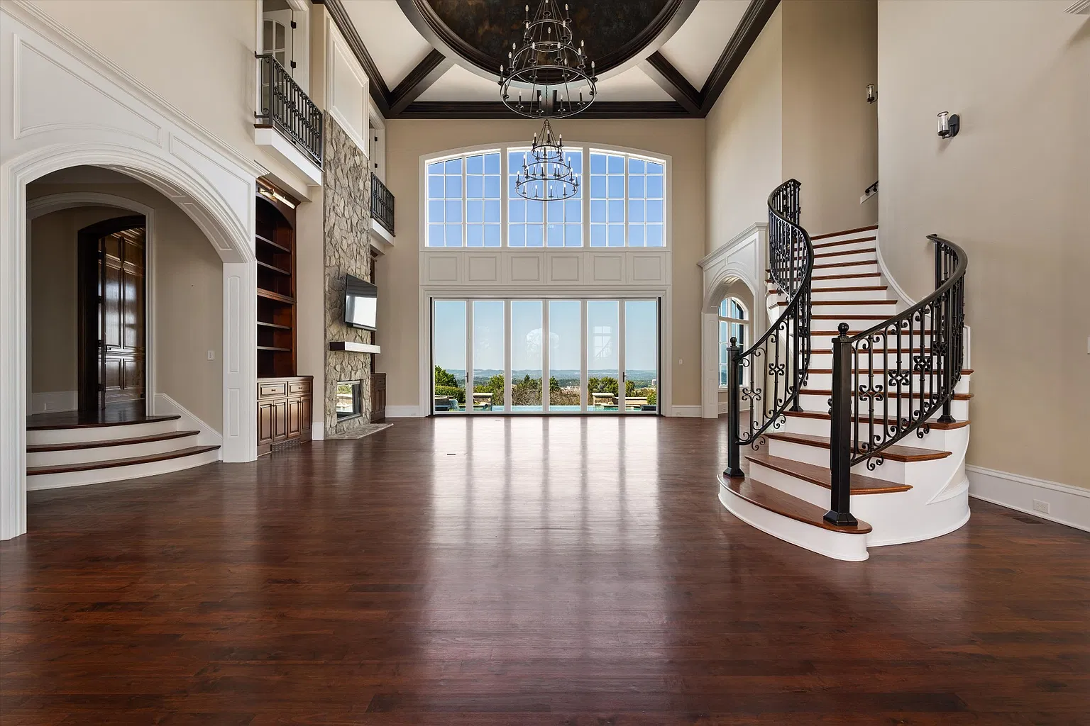 The entire space rises beneath a coffered ceiling with a large circular inset and tiered chandeliers, while a stone fireplace and built-in cabinetry sit on the wall across from the staircase.