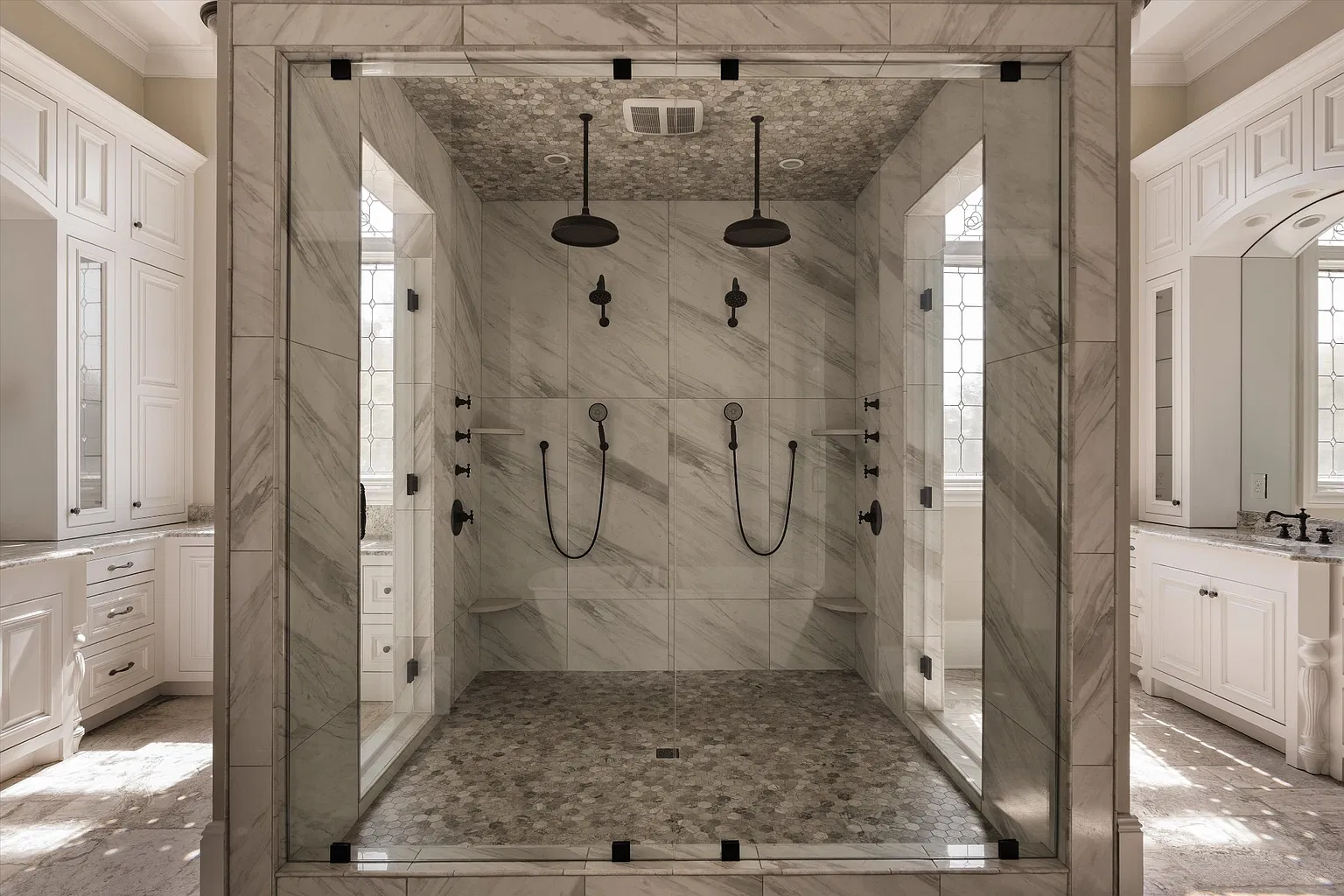 A large glass-enclosed shower lined in veined stone and fitted with dual rainfall heads sits in the middle of matching white vanities.