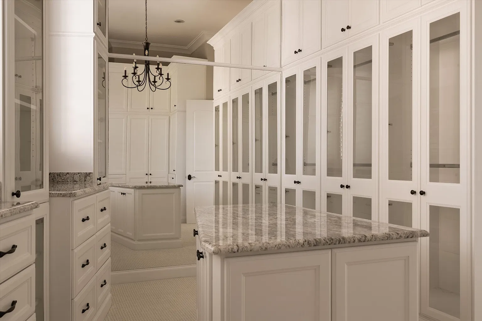 Walk-in closet wrapped in floor-to-ceiling white cabinetry with glass-front doors, drawers, and a central stone-topped island.