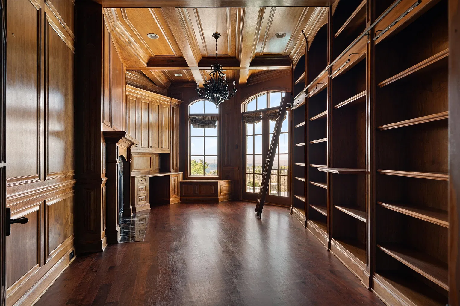 Built-in shelving runs along one wall beneath a coffered wood ceiling, while a fireplace and desk sit opposite tall arched windows and French doors.