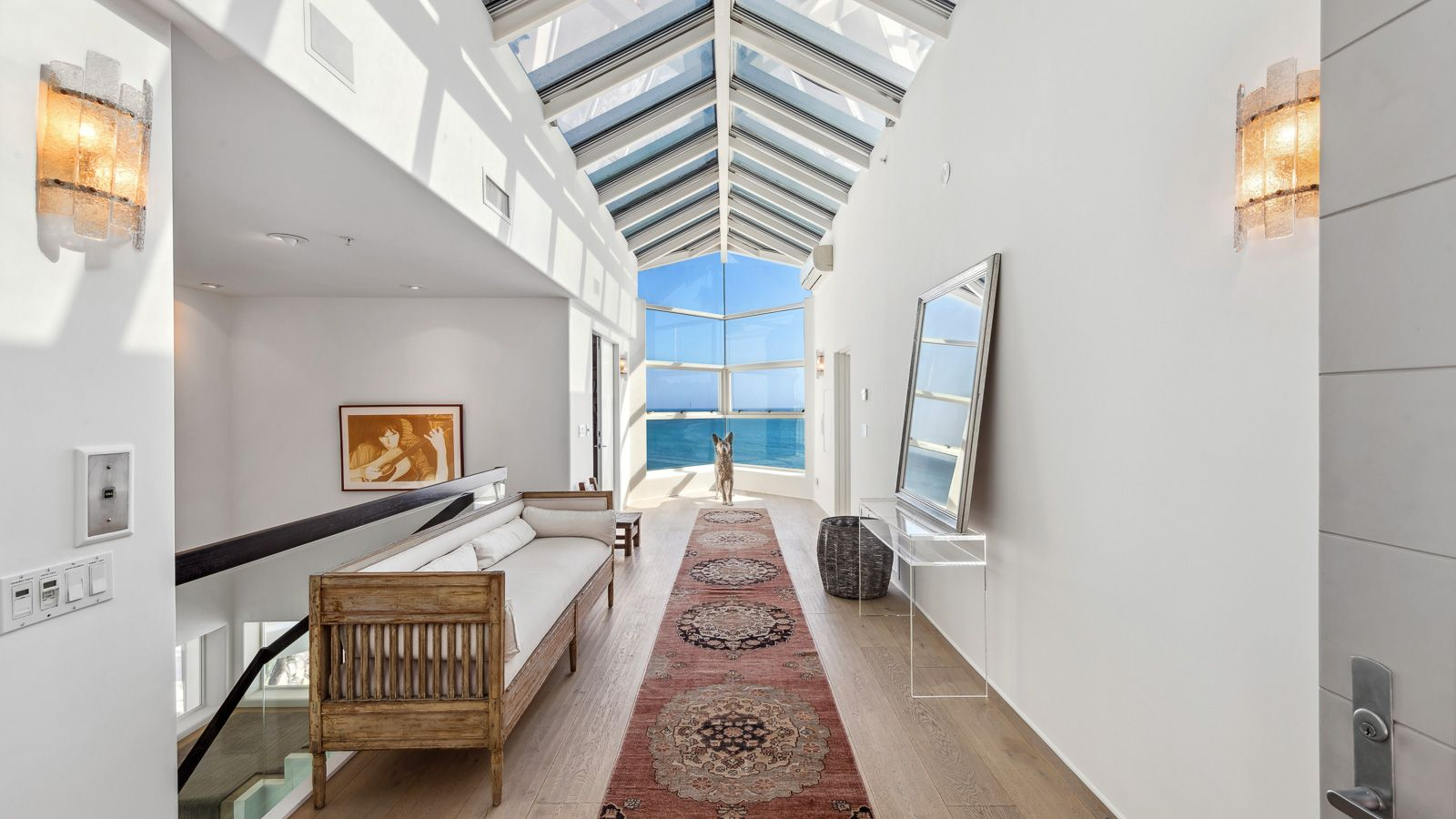 The home opens into a long covered entry hall where vaulted skylights run overhead, and a wall of windows at the far end frames the ocean.