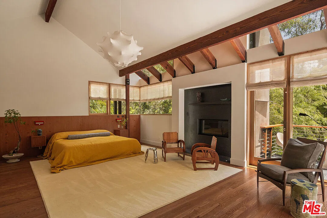 The primary suite's bedroom opens beneath tall, beamed ceilings with the room's windows framing the fireplace wall.