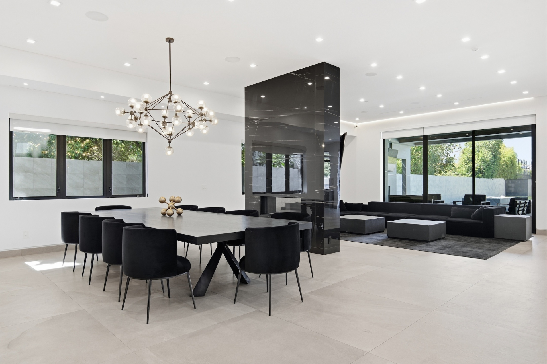 On the other side of the fireplace, the open layout transitions into the dining area, where a long table sits beneath a sculptural chandelier.