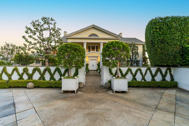 The driveway opens up to reveal the front of the house, framed by geometric hedges, tall sculpted planters, and a central pathway.