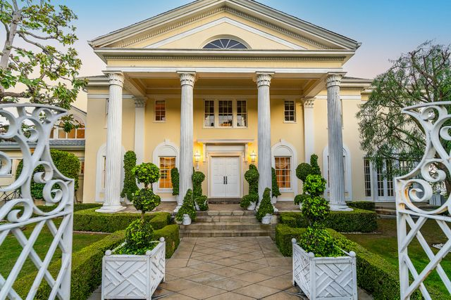 Fluted columns, clipped hedges, and a pair of topiary-lined planters guide you up the steps, where double front doors sit centered beneath the yellow exterior.