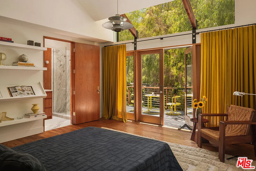 Bedroom framed by exposed beams and upper-level windows, with the bed positioned across from French doors leading to a private balcony.