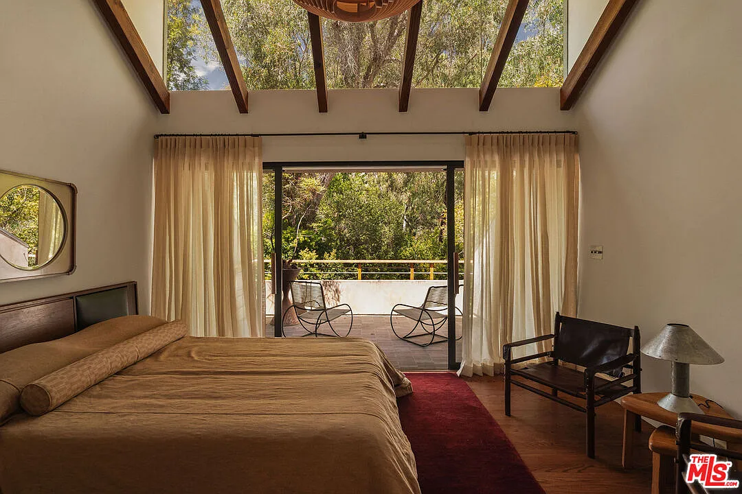The bed set beneath a tall pitched ceiling and a row of exposed beams running overhead to frame a full glass window on the top half of the exterior wall.