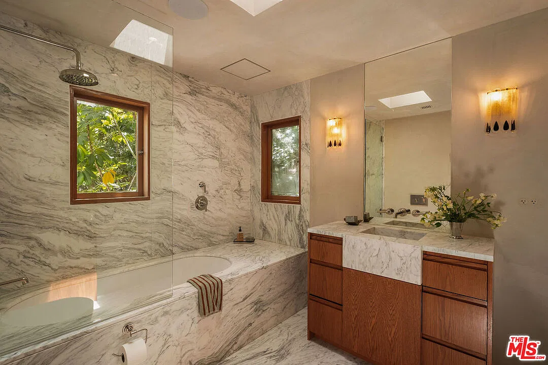 A marble-wrapped soaking tub placed beneath two small windows, with a wood vanity next to it and a skylight overhead.
