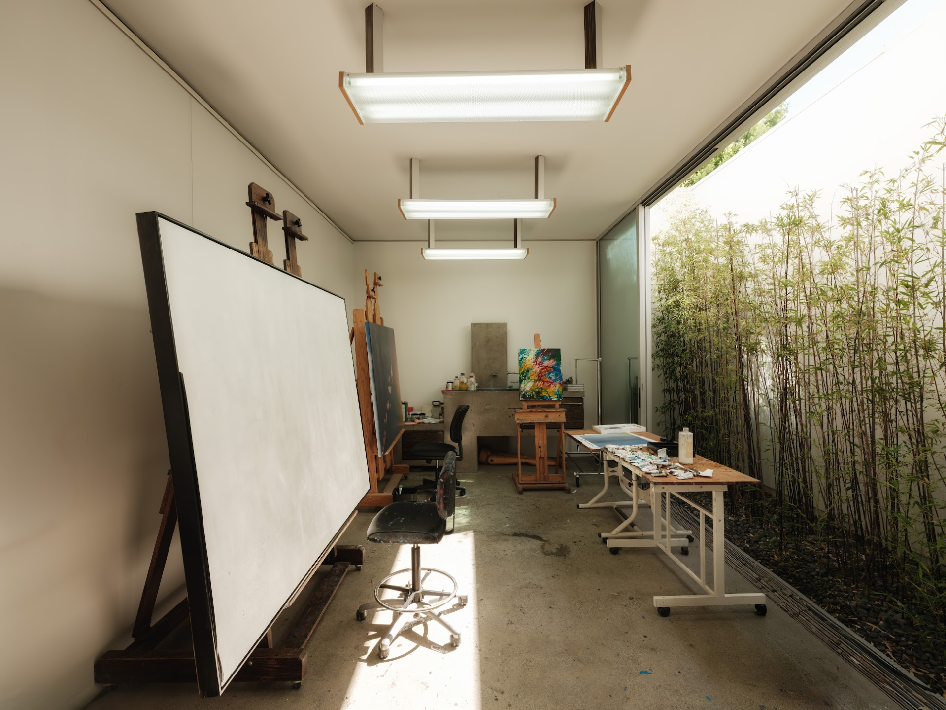 A long, narrow room lined with easels and worktables, with a full wall of glass opening toward a bamboo-lined garden.