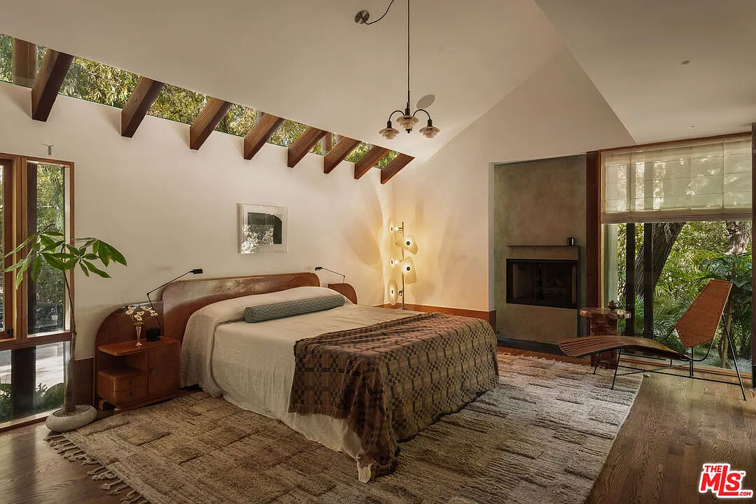 The bed sits beneath a high sloped ceiling, while a fireplace occupies the far wall beside glass doors that open to the backyard.