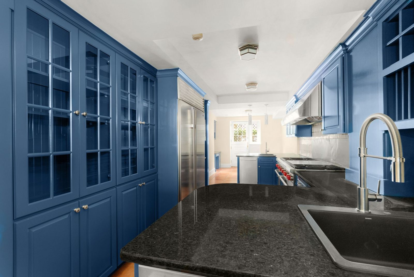 Blue cabinetry lines both sides of the room around black countertops, with tall storage and refrigeration built into one wall and a large range set beneath a stainless-steel hood on the other.