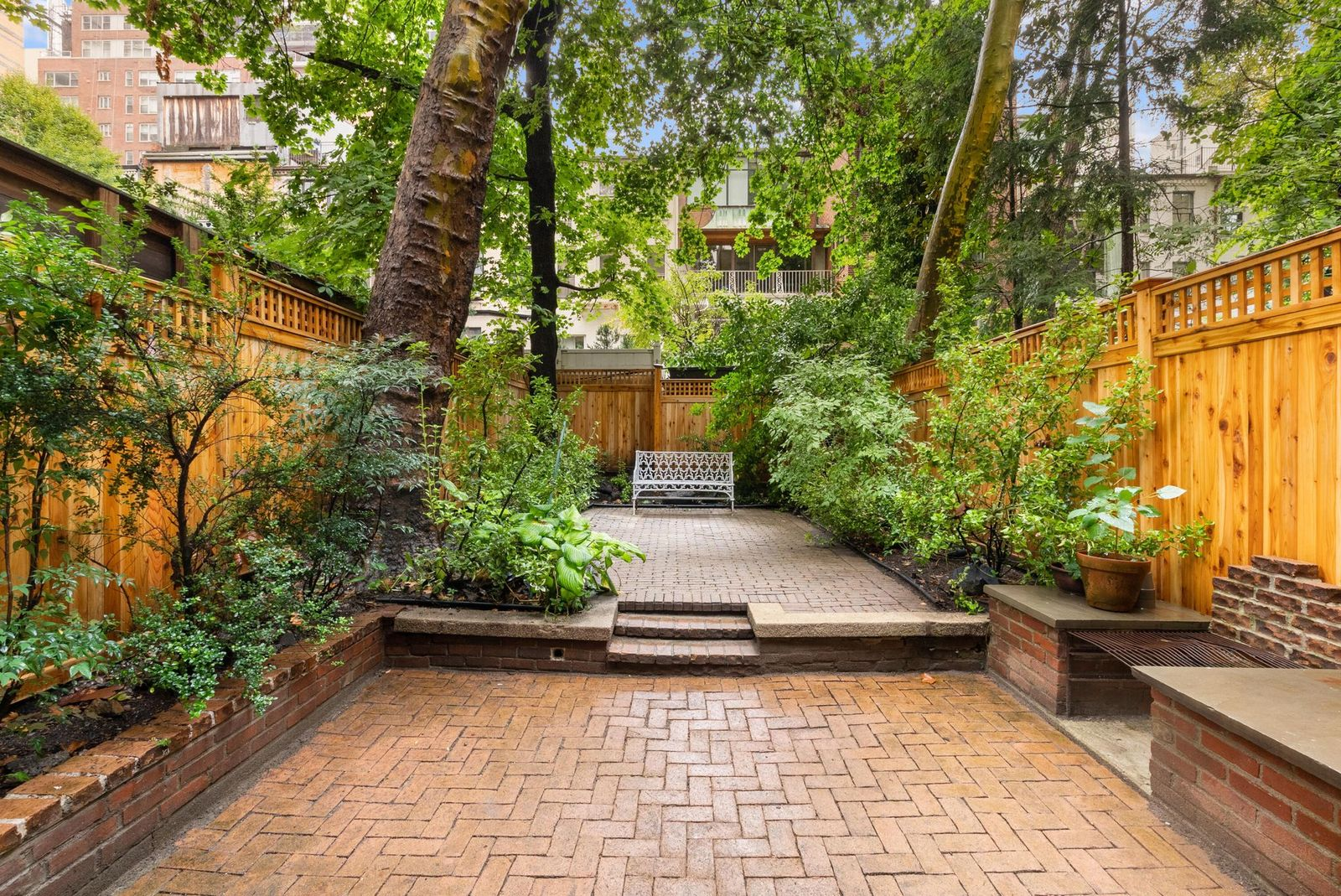 The outdoor space has two levels of brick patios, with the lower terrace stepping up to a second paved area surrounded by planting beds and trees.