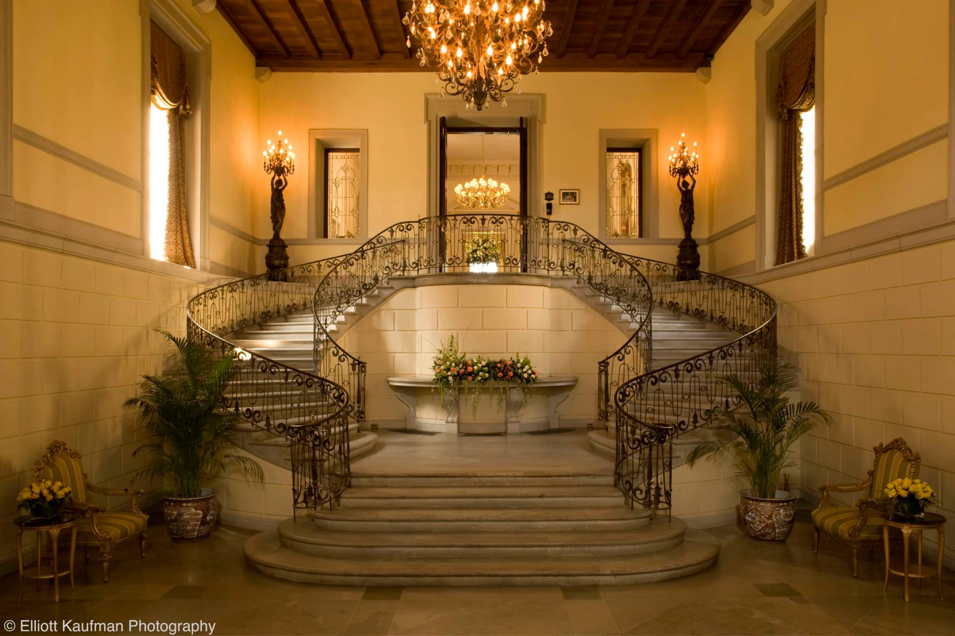 Two sweeping flights curve up from the main hall beneath a coffered wood ceiling, with wrought-iron railings tracing the stairs.
