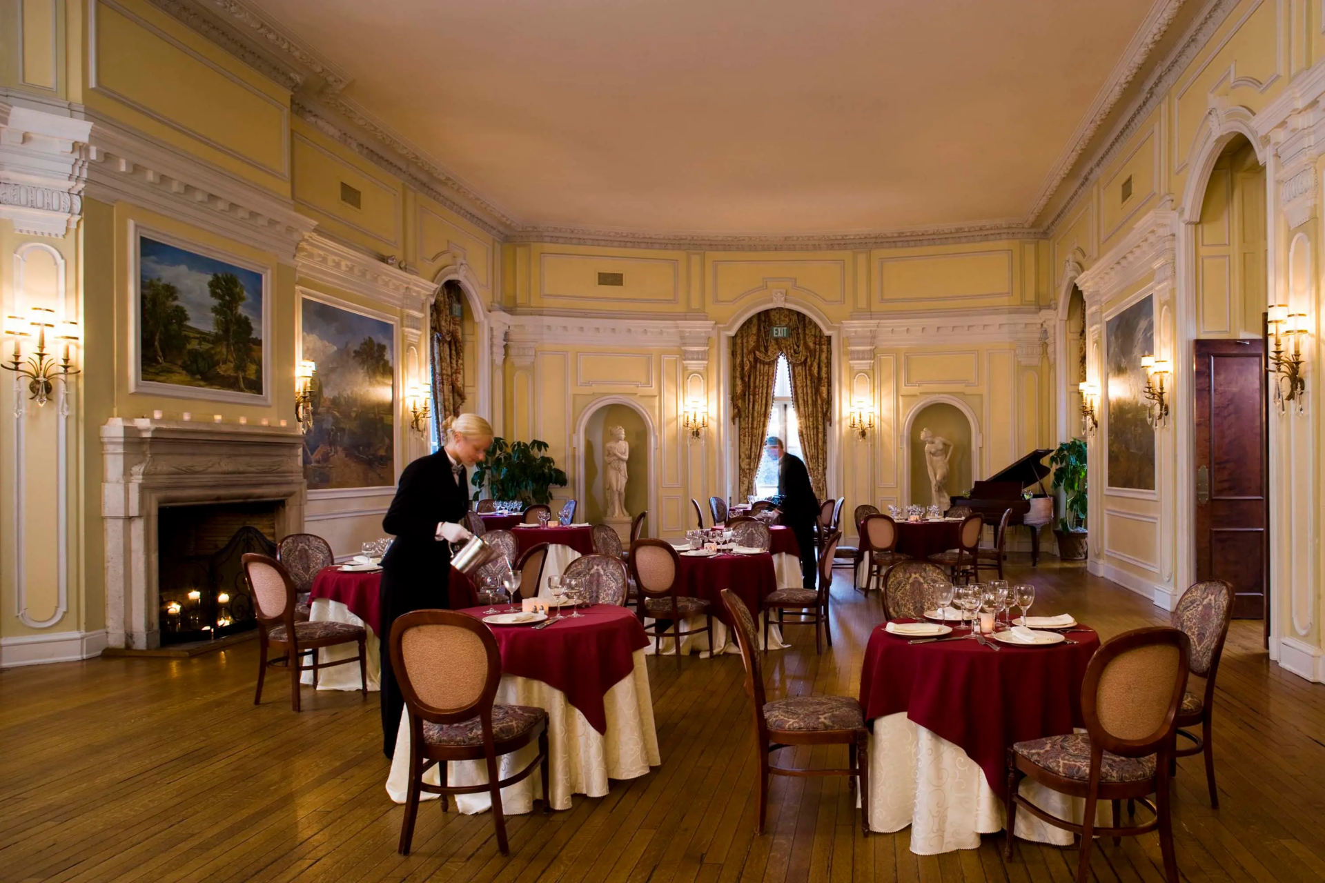 Round dining tables are spread across the floor beneath a high ceiling, while statues and framed landscape paintings fill the room.