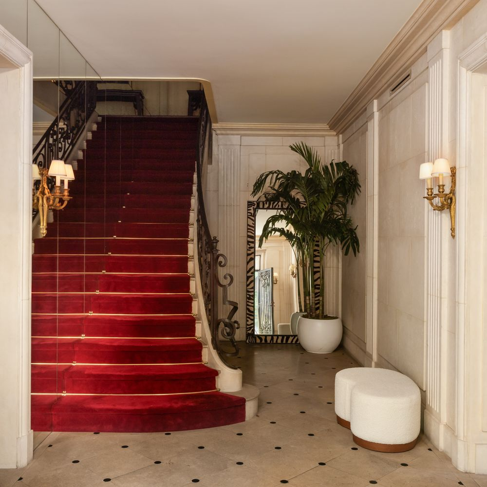 Off to one side, a red-carpeted staircase rises next to a mirrored wall, while pale stone and brass sconces line the rest of the walls.