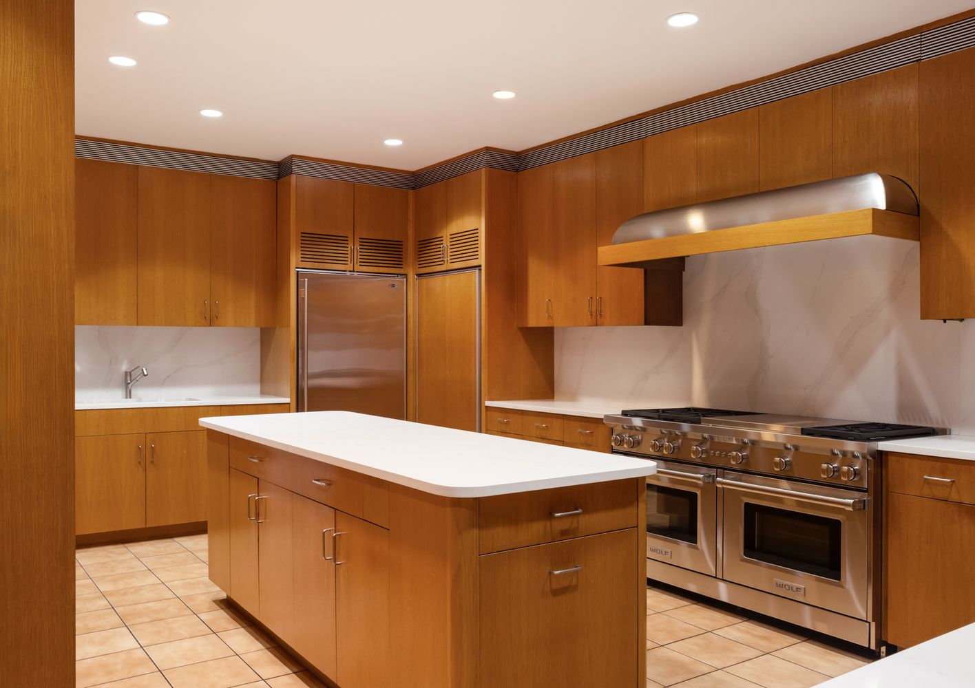 The kitchen is organized around a long central island, with white counters running between upper and lower wood cabinetry. 