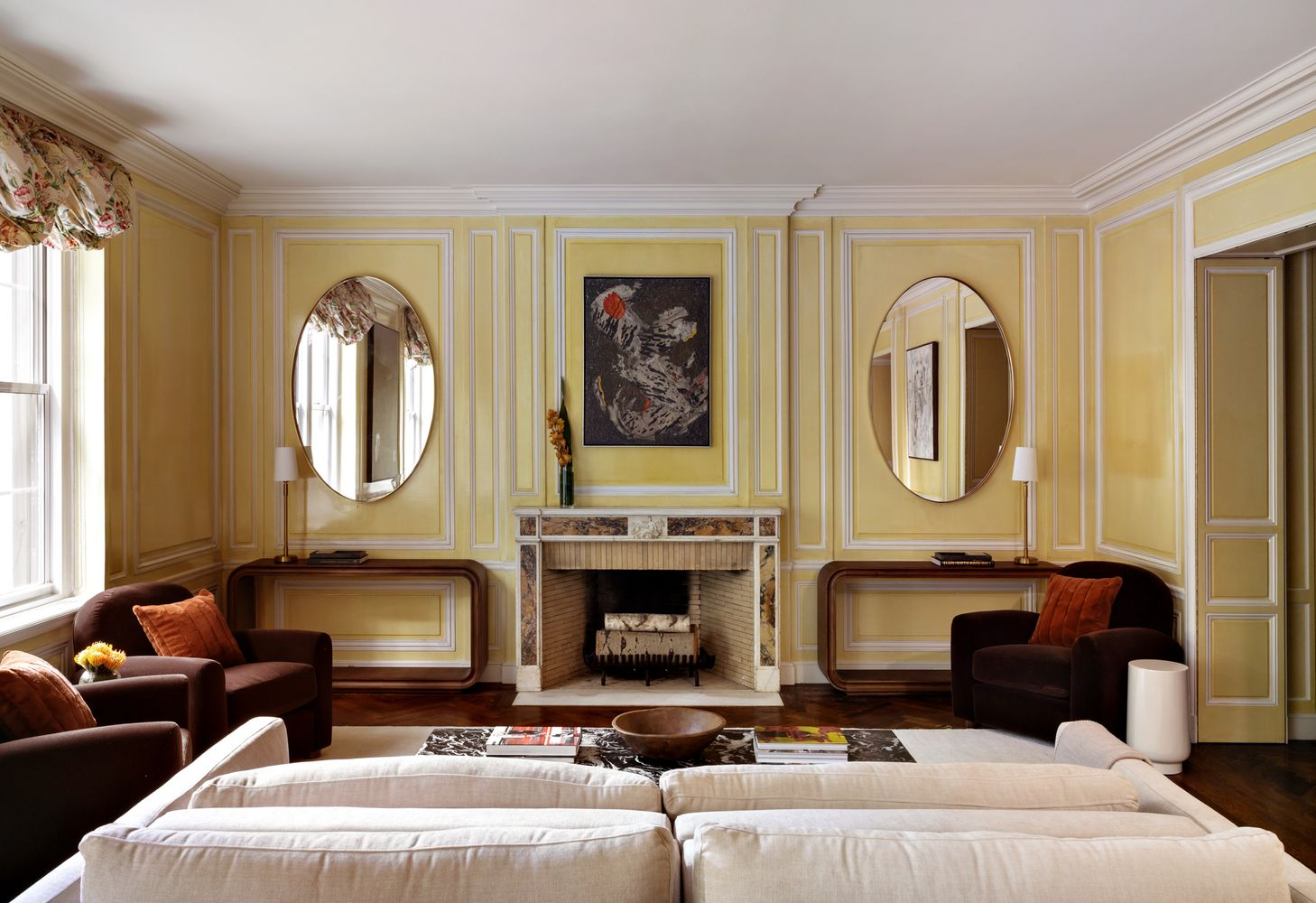 Pale yellow paneled walls wrap the room with matching console tables and oval mirrors placed on either side of the fireplace.