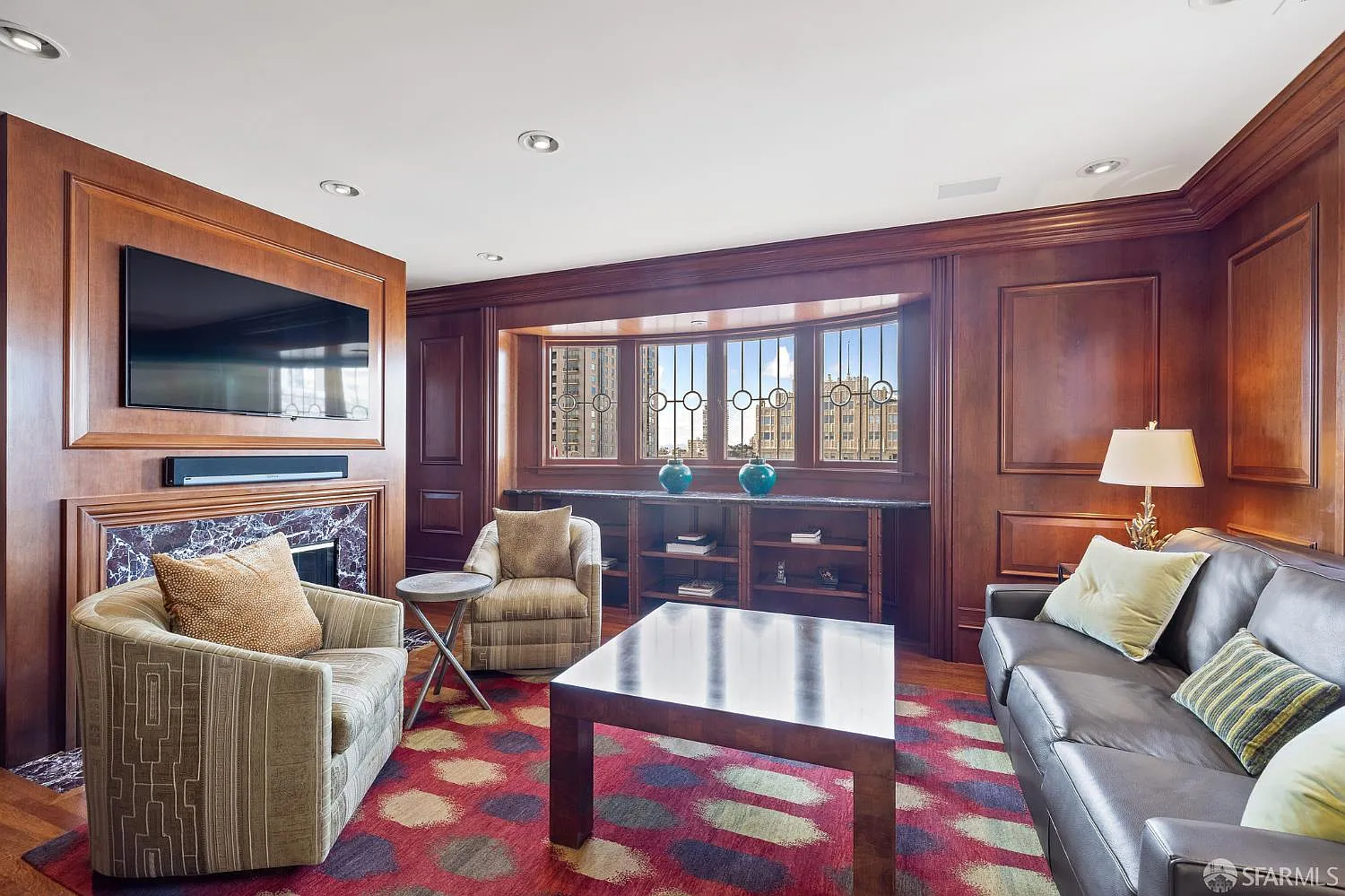 Dark wood wraps the walls around a deep window bay while a leather sofa and two lounge chairs sit across from a marble fireplace and mounted TV.